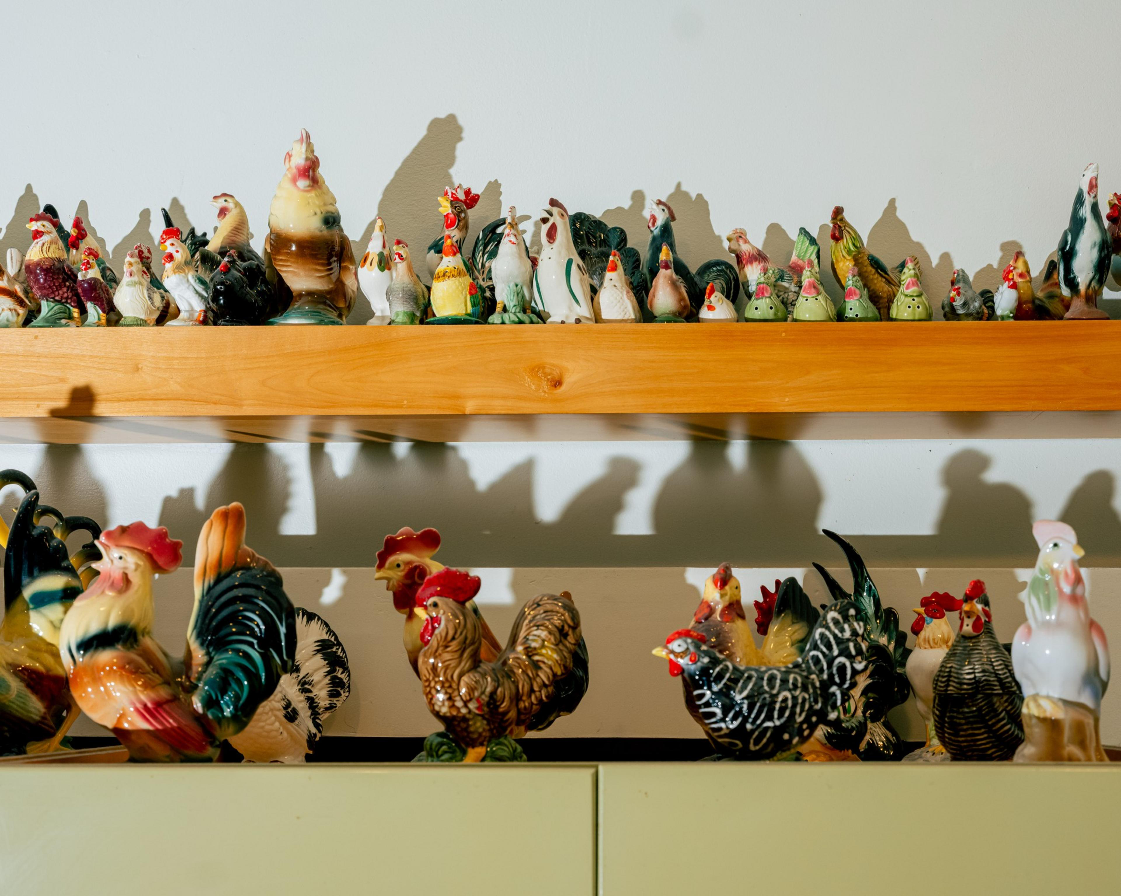 Two shelves display numerous ceramic rooster and chicken figurines in various colors, sizes, and detailed designs against a white wall.
