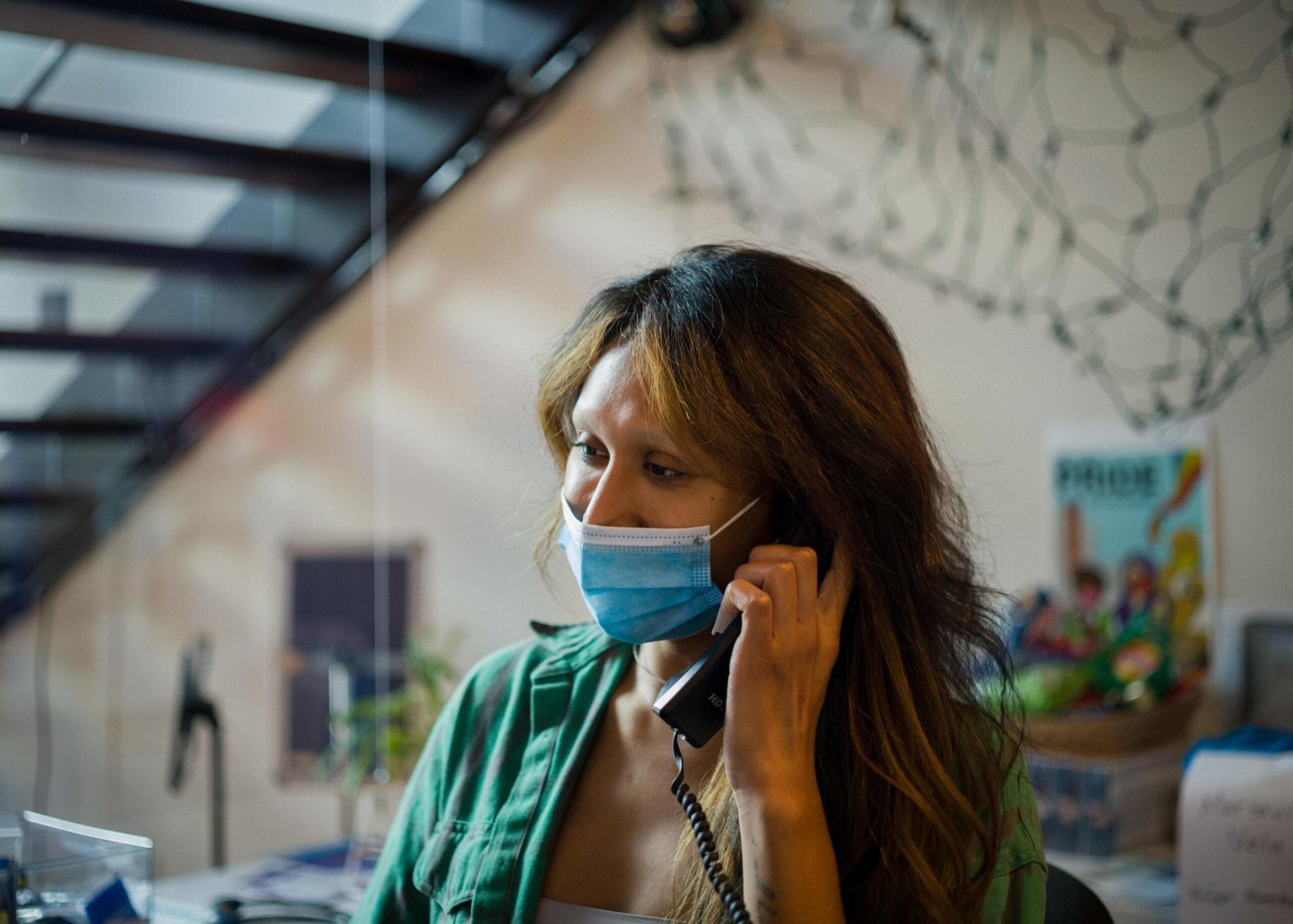 A woman wearing a blue face mask and green shirt talks on a corded phone indoors with a staircase and colorful poster in the background.