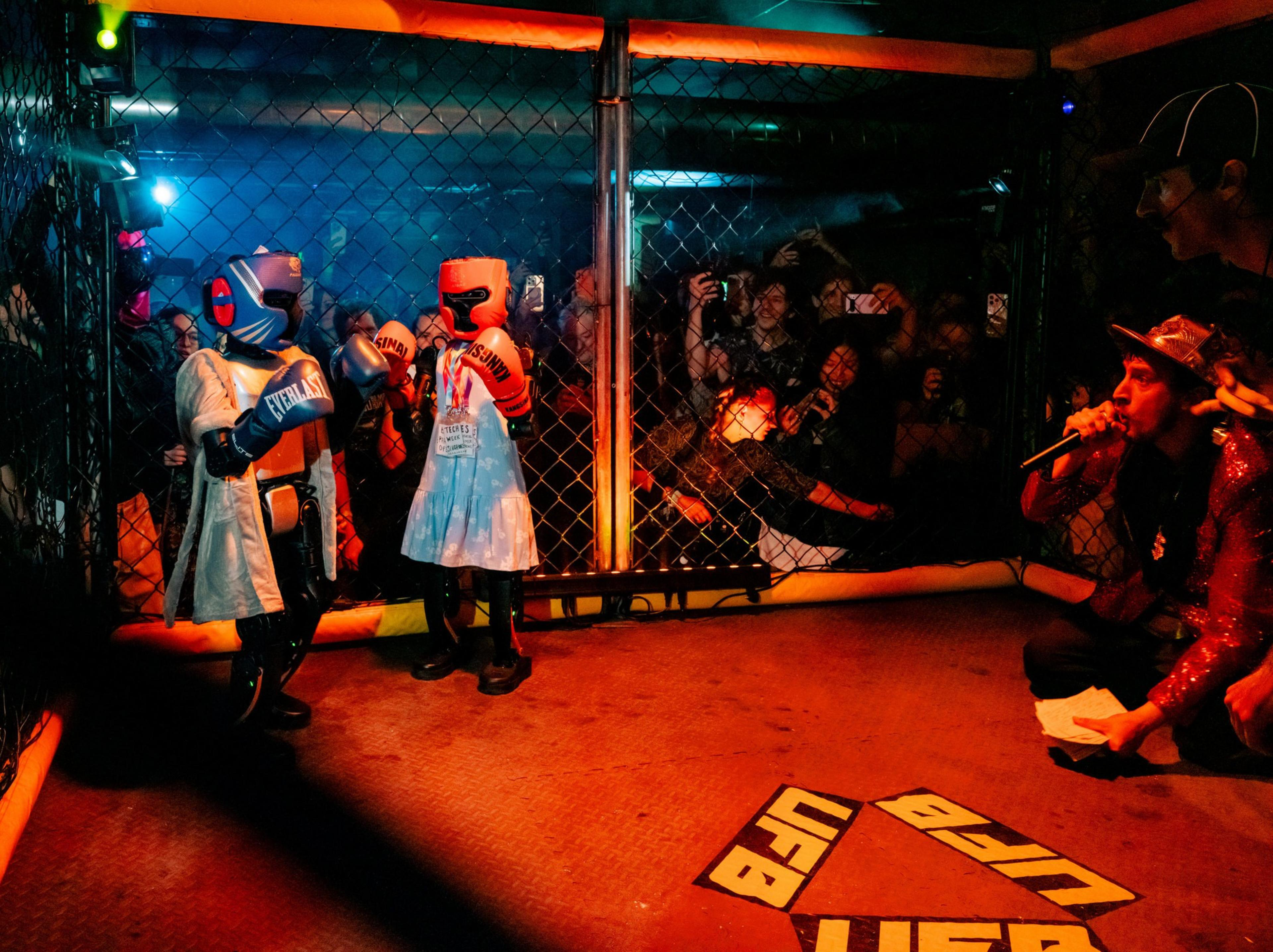 Two young children in boxing gear face each other inside a cage while an announcer speaks into a microphone nearby.
