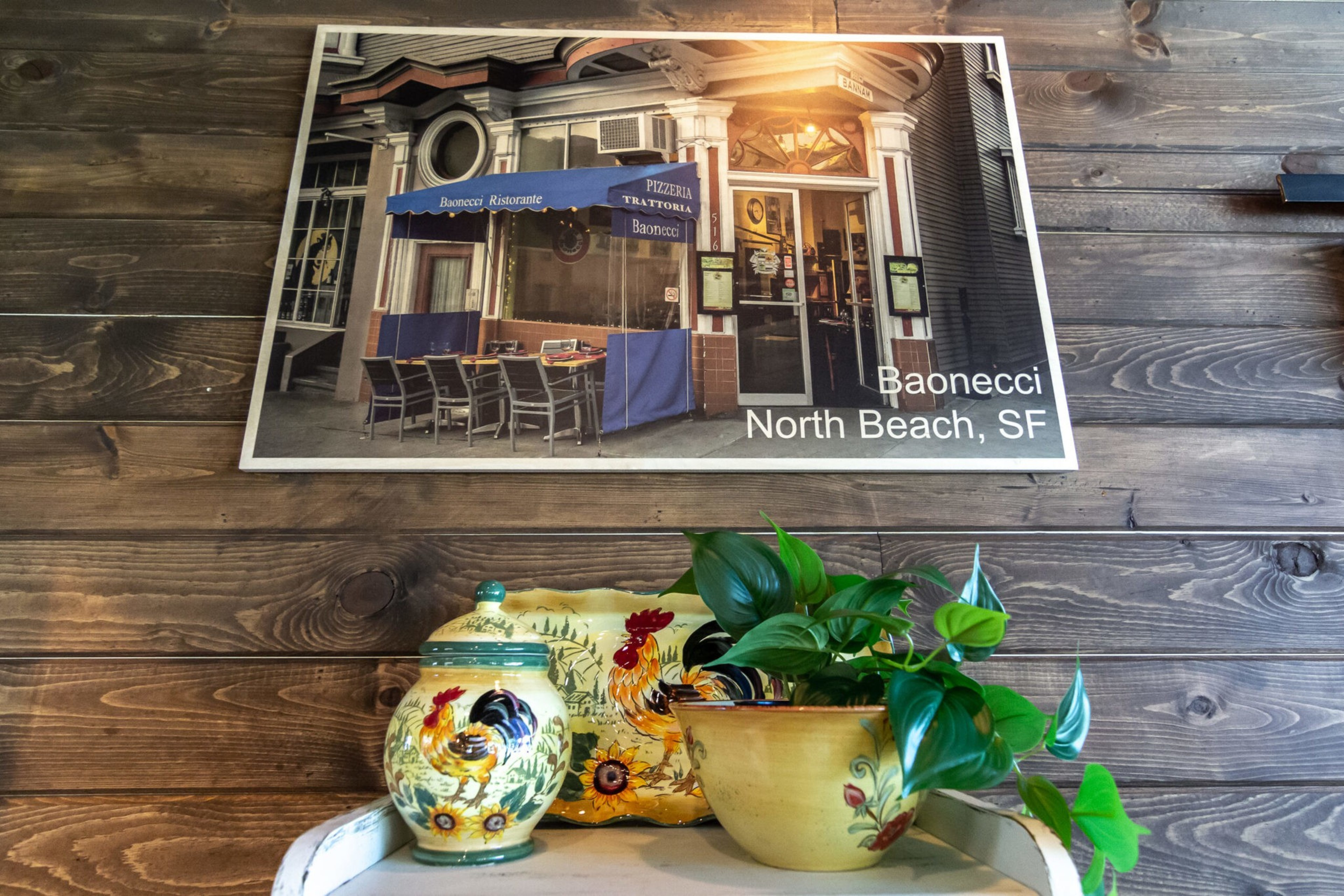 A framed photo of Baonecci restaurant in San Francisco hangs on a wooden wall above ceramic containers and a green leafy plant.