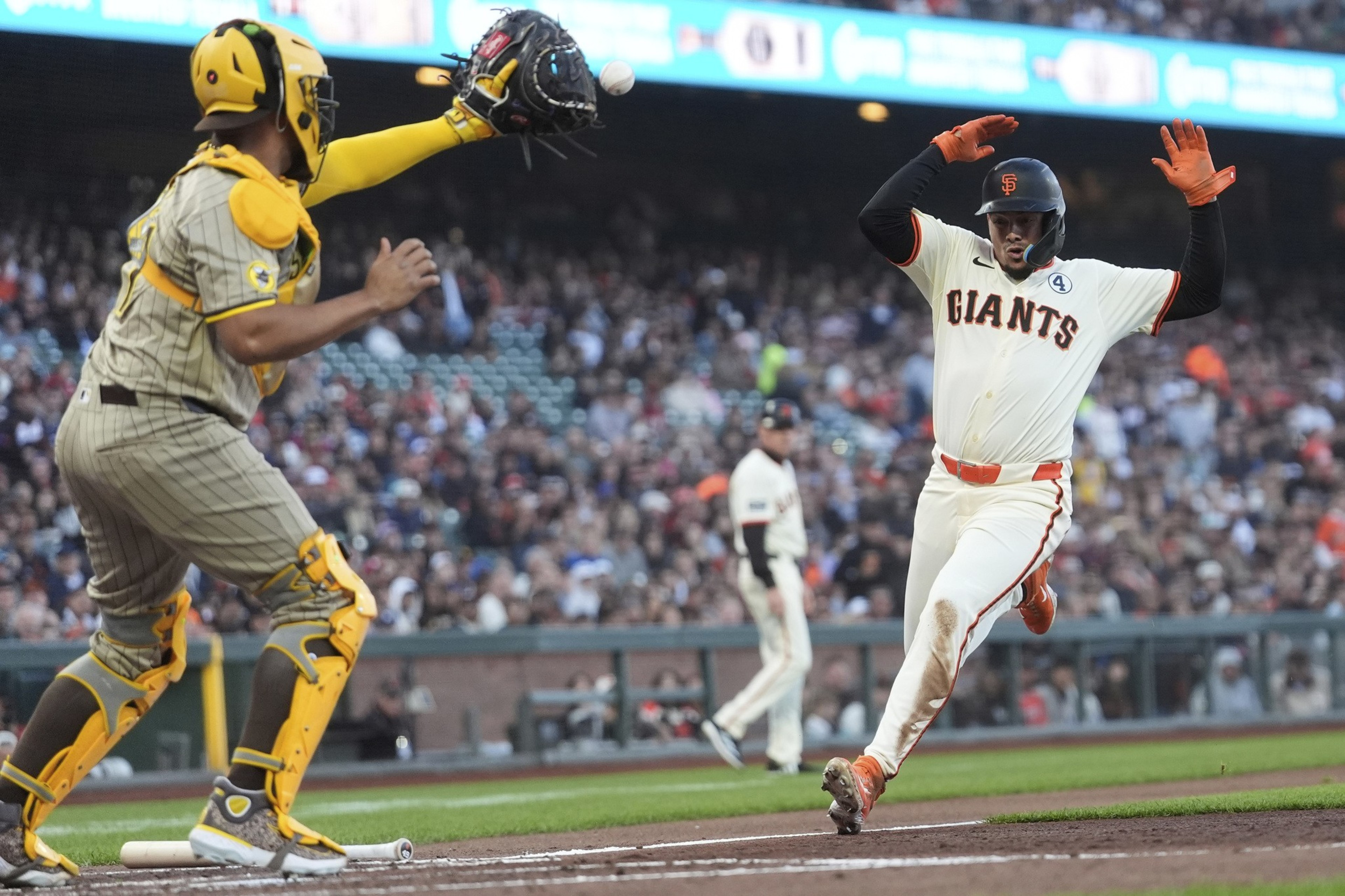 A baseball player from the Giants slides toward home plate as a catcher in yellow gear reaches with his glove to tag him, with spectators in the background.