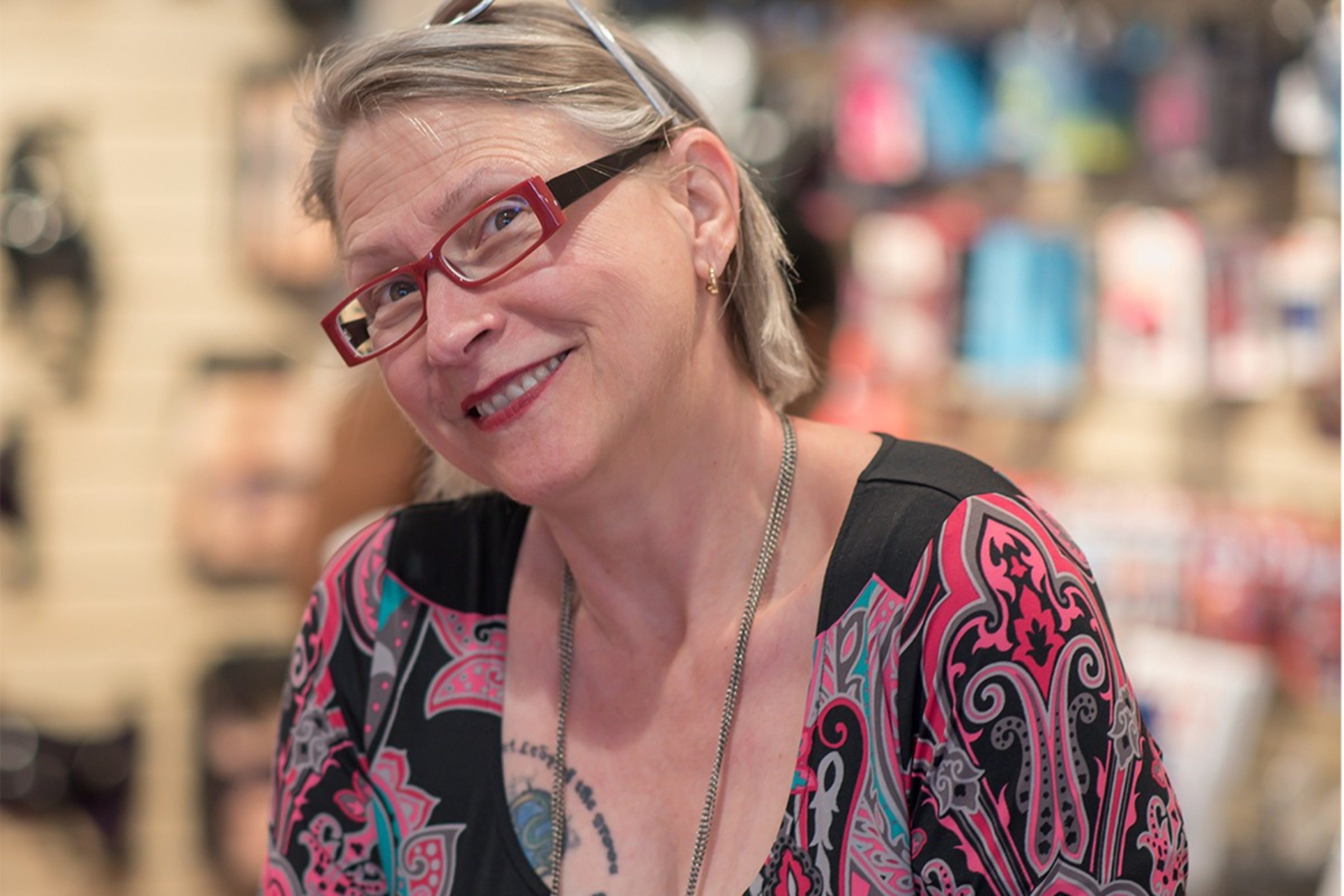 A woman with short hair and red glasses smiles at the camera. She wears a colorful patterned top and a necklace, with blurred shelves in the background.