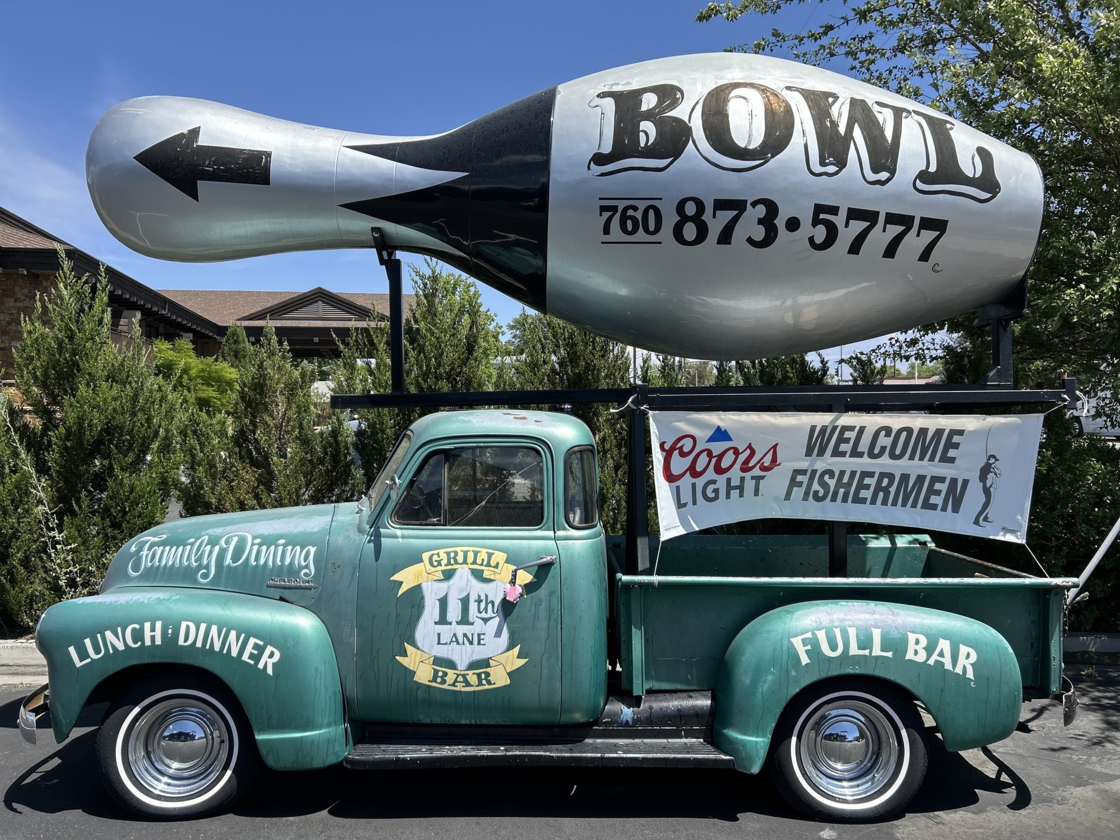 A vintage green truck is parked with a large bowling pin sign on top, advertising &quot;BOWL&quot; and a phone number. The truck features &quot;Family Dining,&quot; &quot;Grill 11th Lane Bar,&quot; and a banner saying &quot;Coors Light, Welcome Fishermen.&quot;