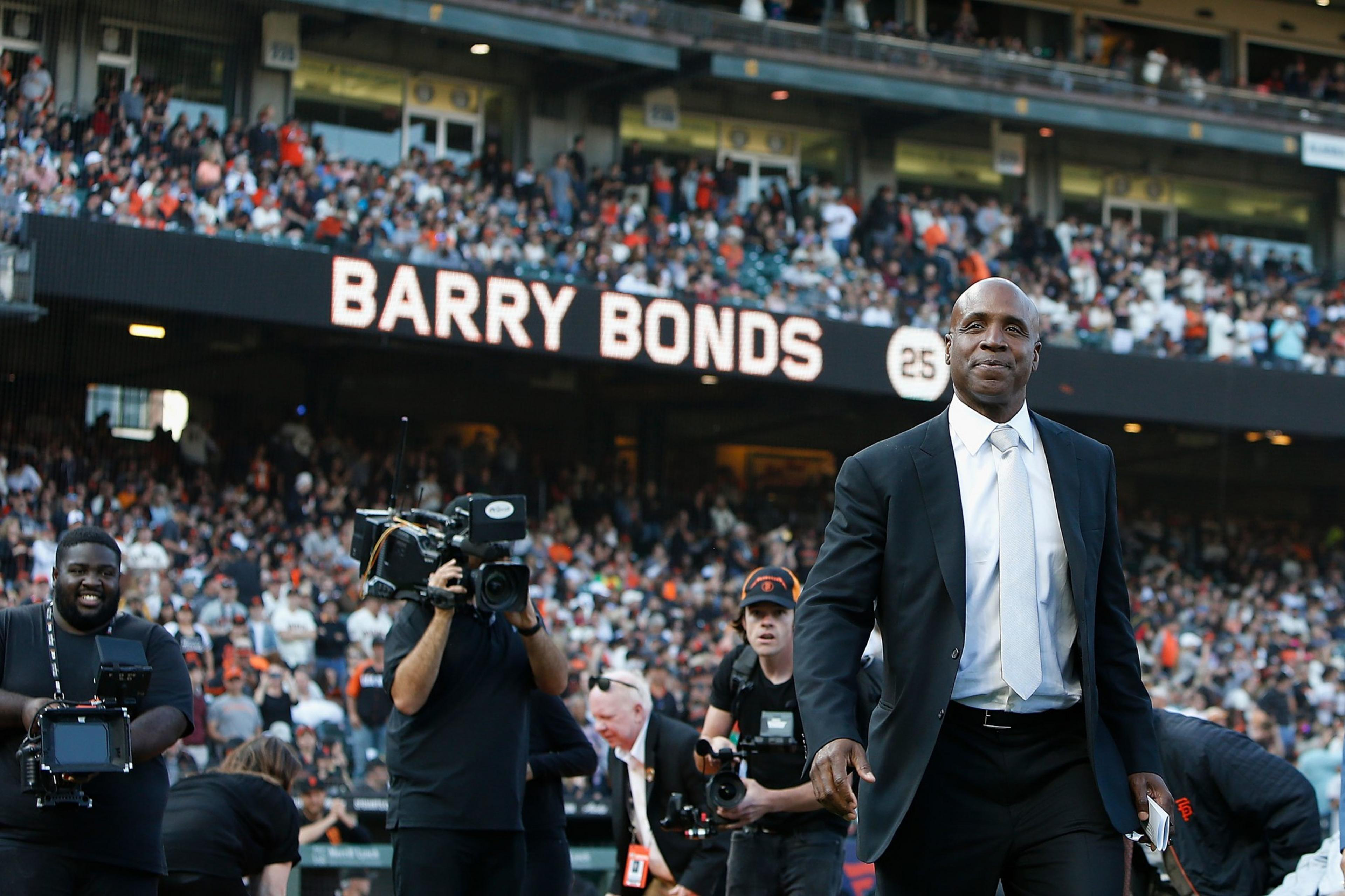 Barry Bonds in a black suit and white tie stands on a baseball field with a large crowd behind him and his name displayed on a scoreboard.