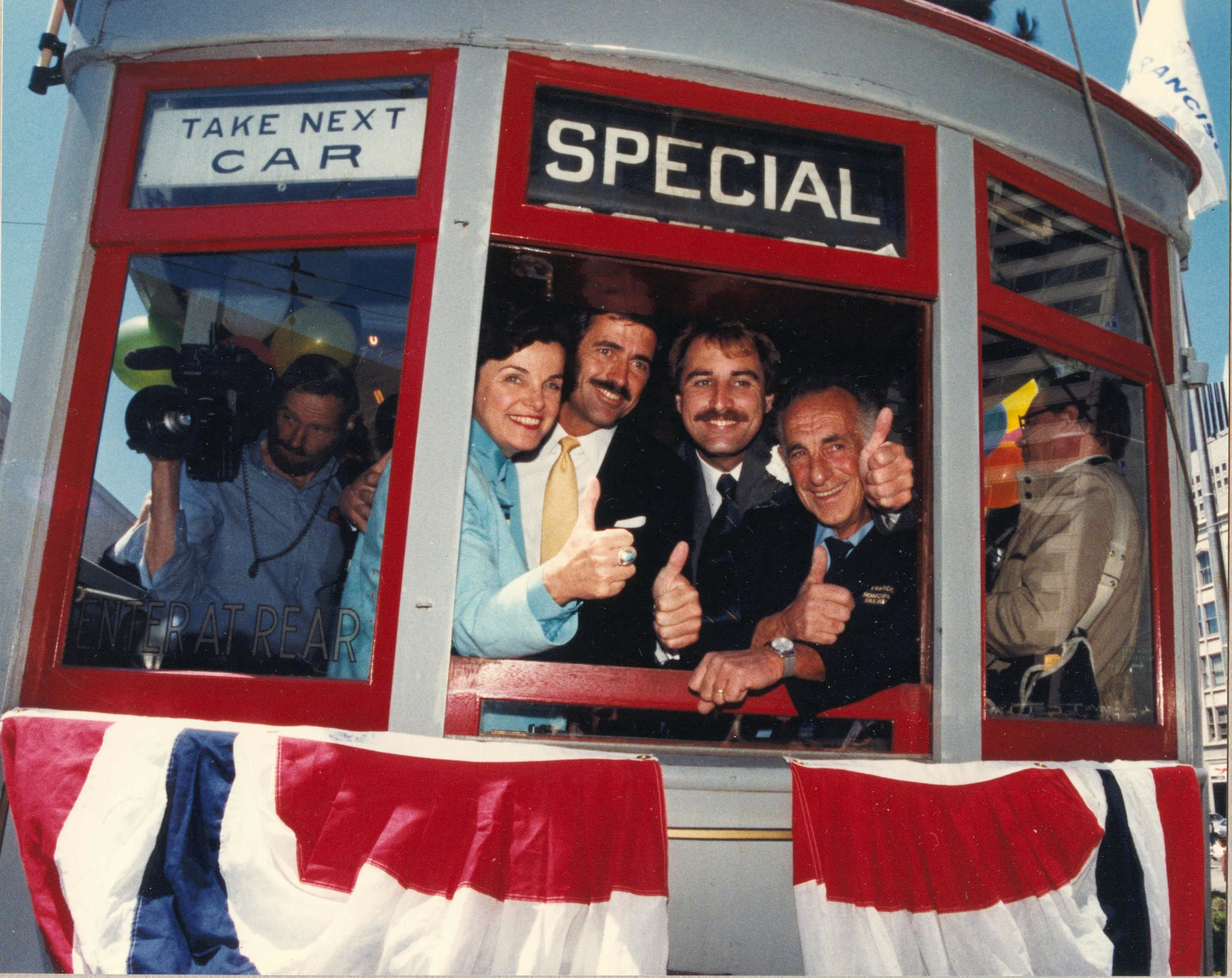 Four people inside a streetcar labeled "SPECIAL" give thumbs up, while a cameraman films through a side window, and red, white, and blue bunting decorates the exterior.
