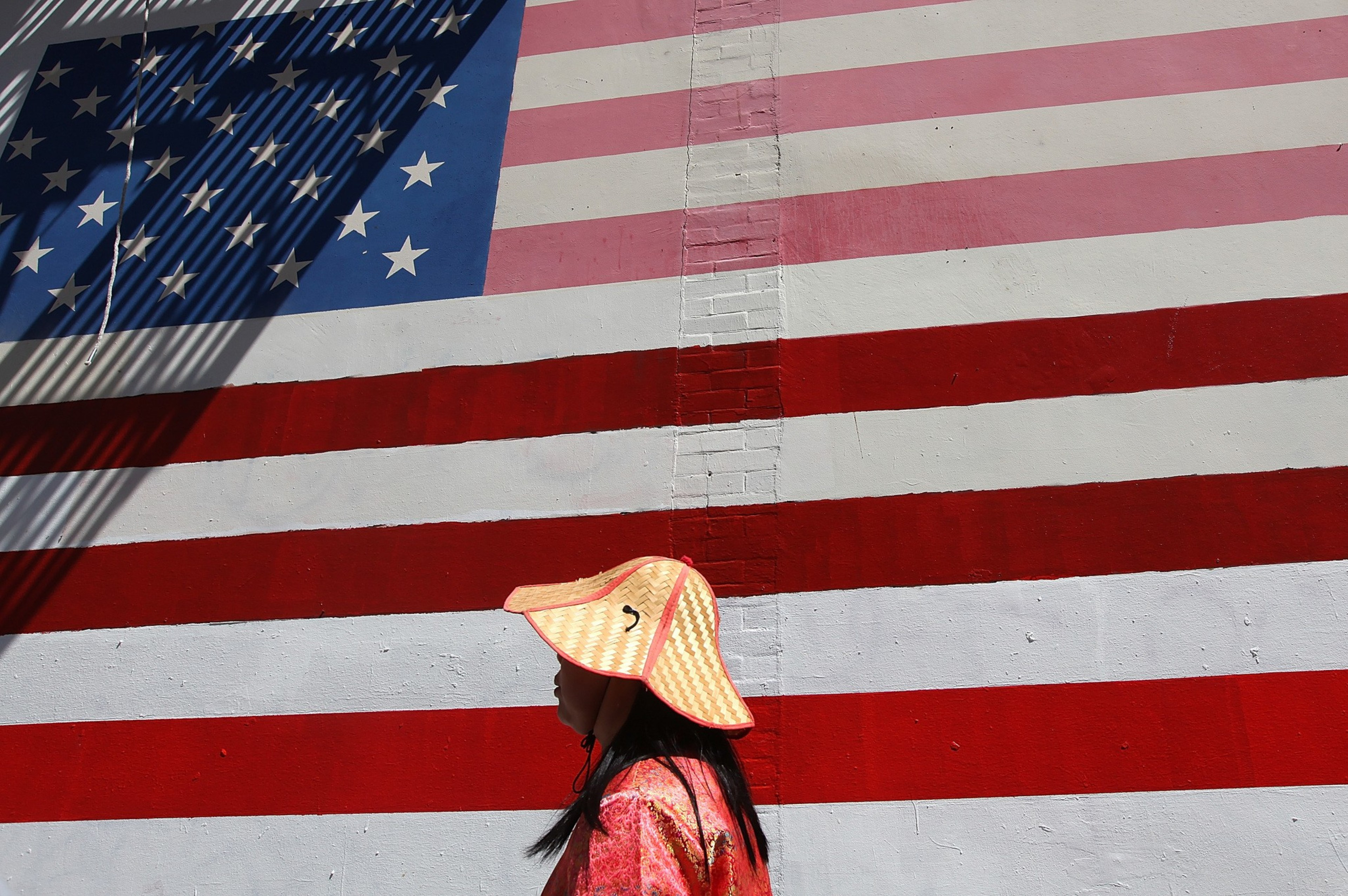 A person in a straw hat walks past a wall painted with the American flag, casting shadows that add texture to the scene.