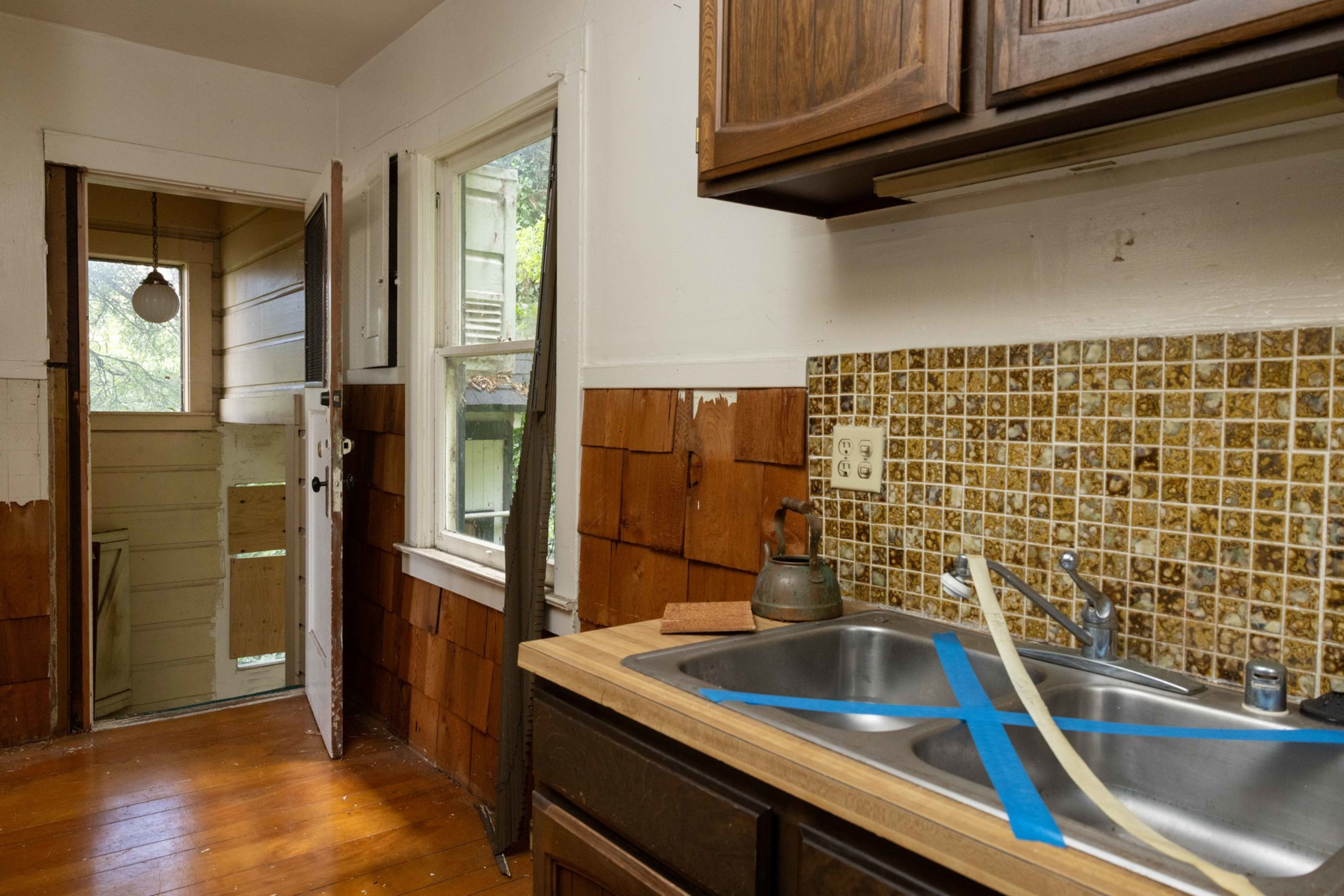 A kitchen with wooden cabinets, a double sink taped off with blue tape, a tiled backsplash, a kettle on the counter, and a door leading to an enclosed porch.
