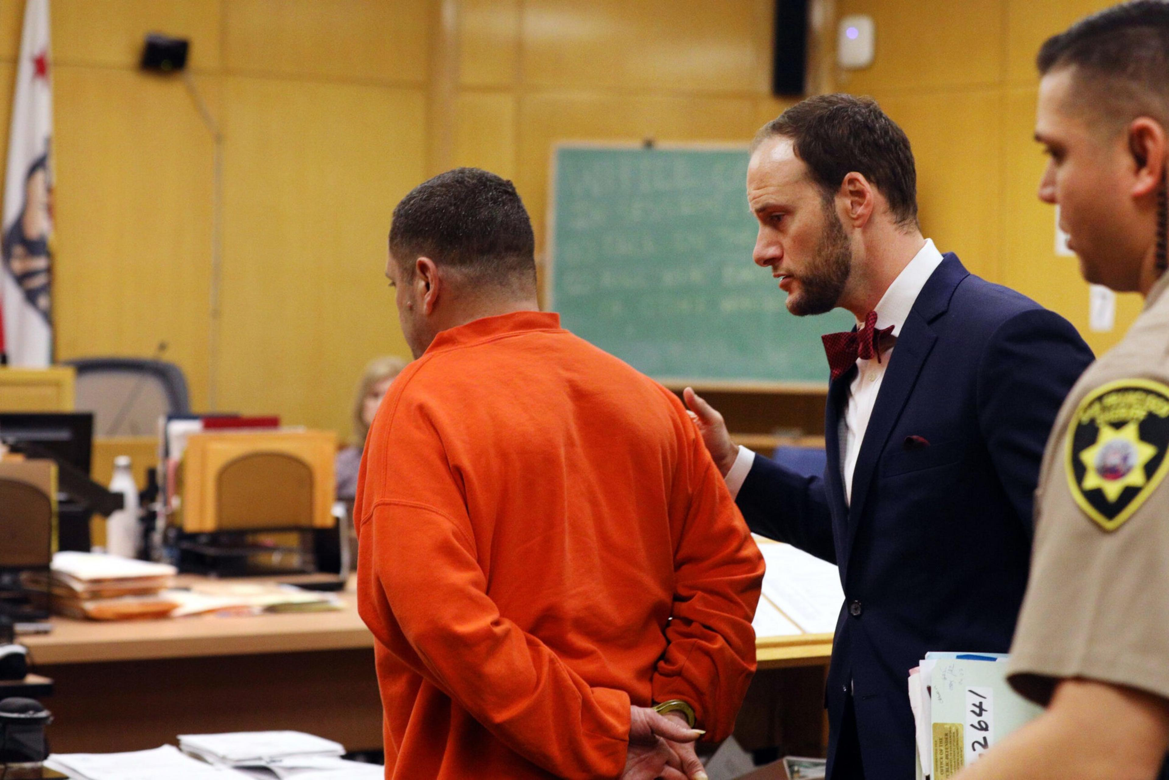 A man in an orange jumpsuit with handcuffs is being spoken to by a man in a suit, while a uniformed officer stands nearby in a courtroom.