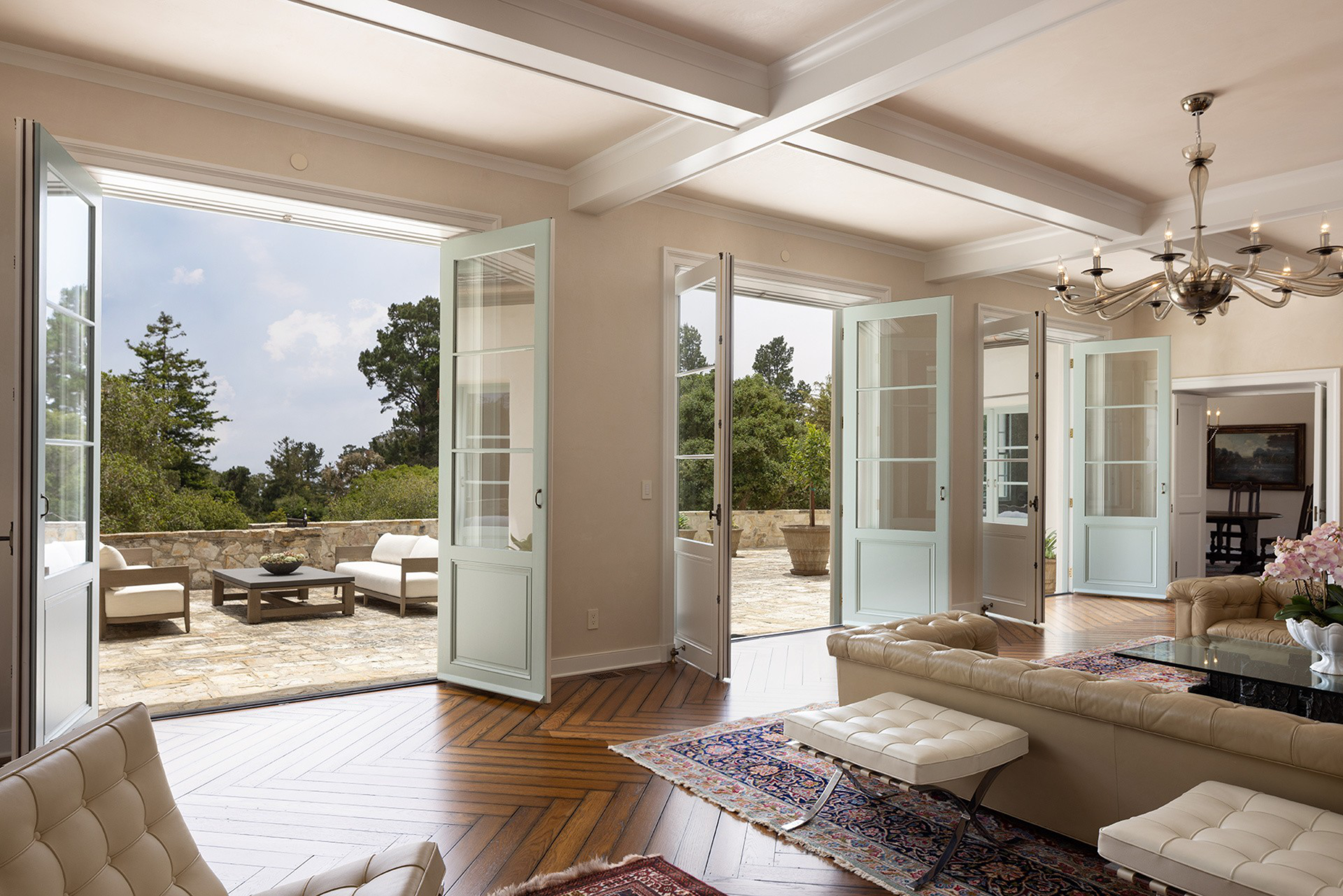 A bright living room with multiple open glass doors leading to a stone patio with outdoor seating, featuring beige leather furniture and wooden floors.