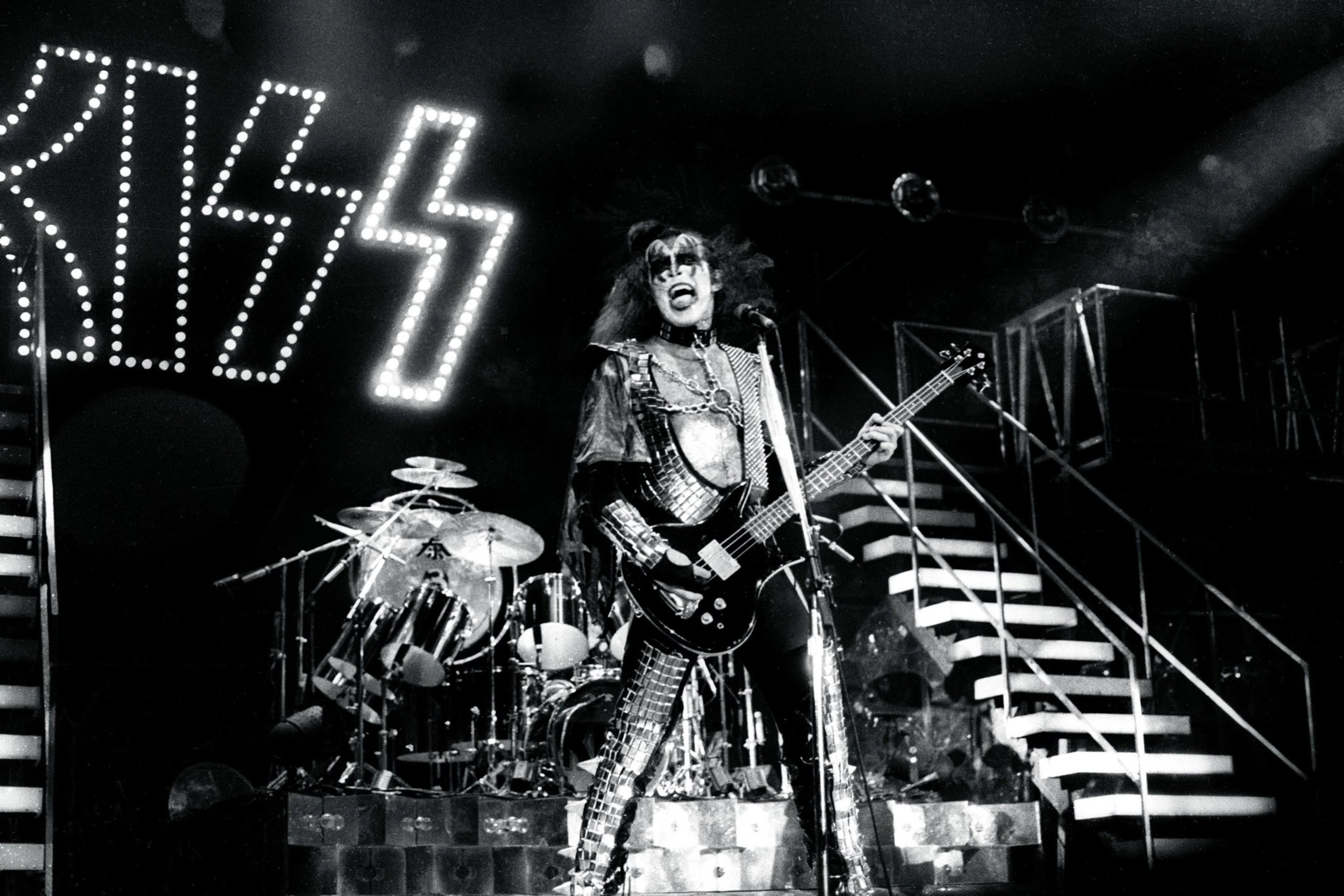 A musician in dramatic makeup and costume plays guitar onstage with &quot;KISS&quot; in lights behind.