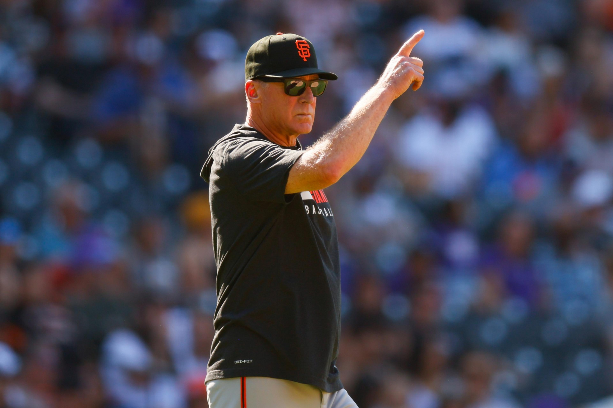 A man wearing a San Francisco Giants cap and black shirt raises his right arm, pointing with his index finger during a baseball game.