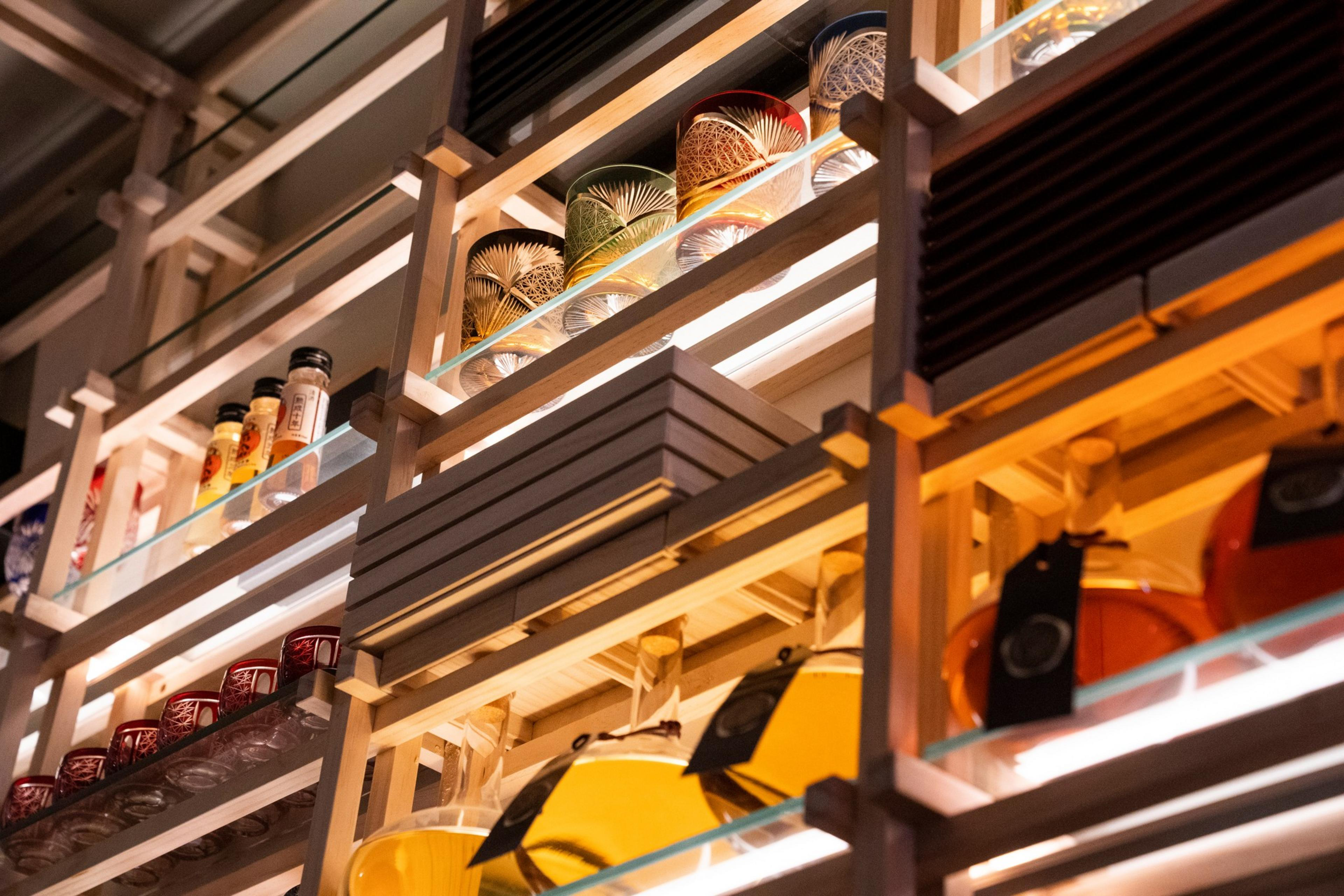 The image shows wooden shelves displaying various glass items, bottles, and decorative objects. The shelves are well-lit, showcasing intricate designs and vibrant colors.