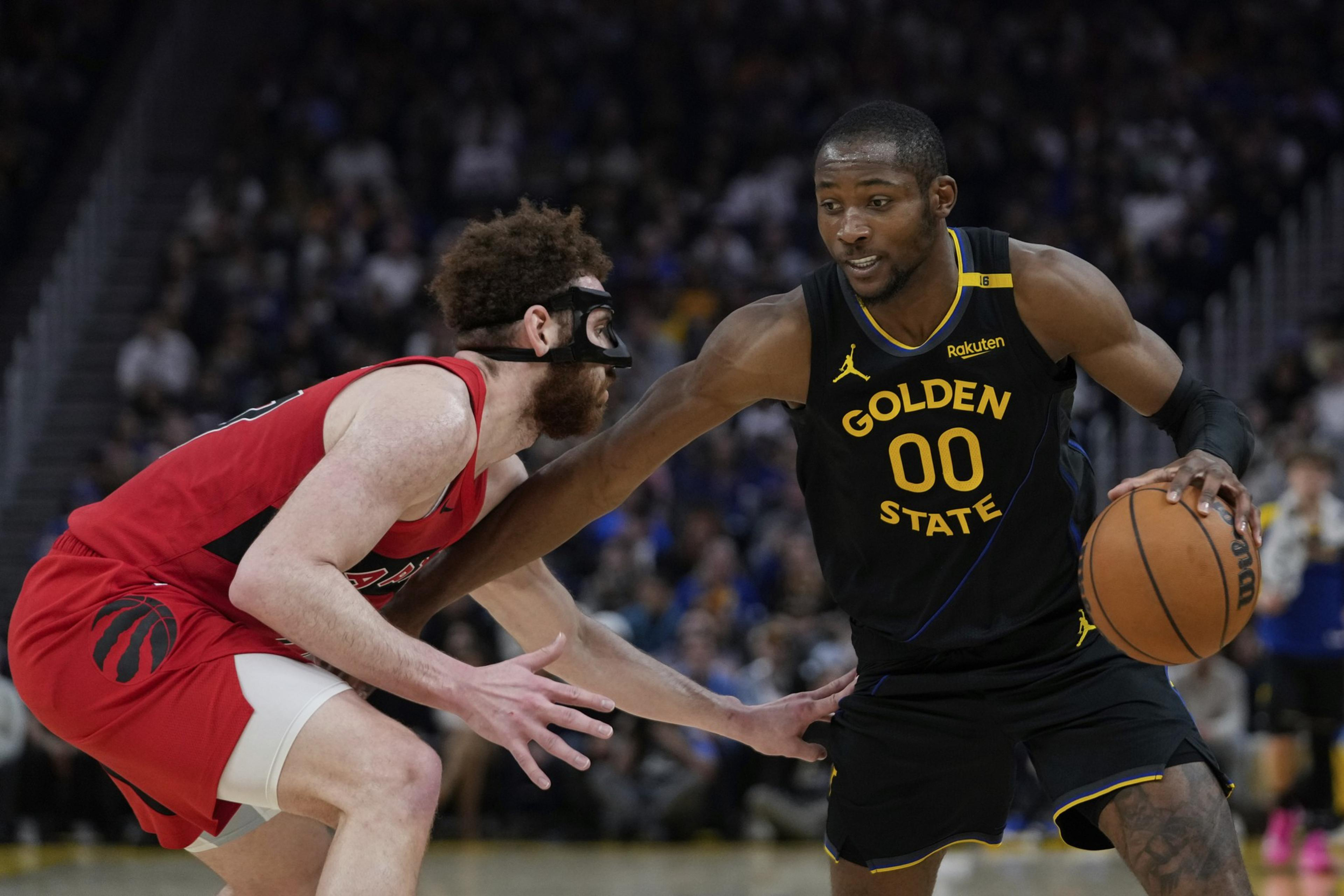 A basketball player in a Golden State uniform dribbles while being guarded by a player in a red uniform with a black face mask.