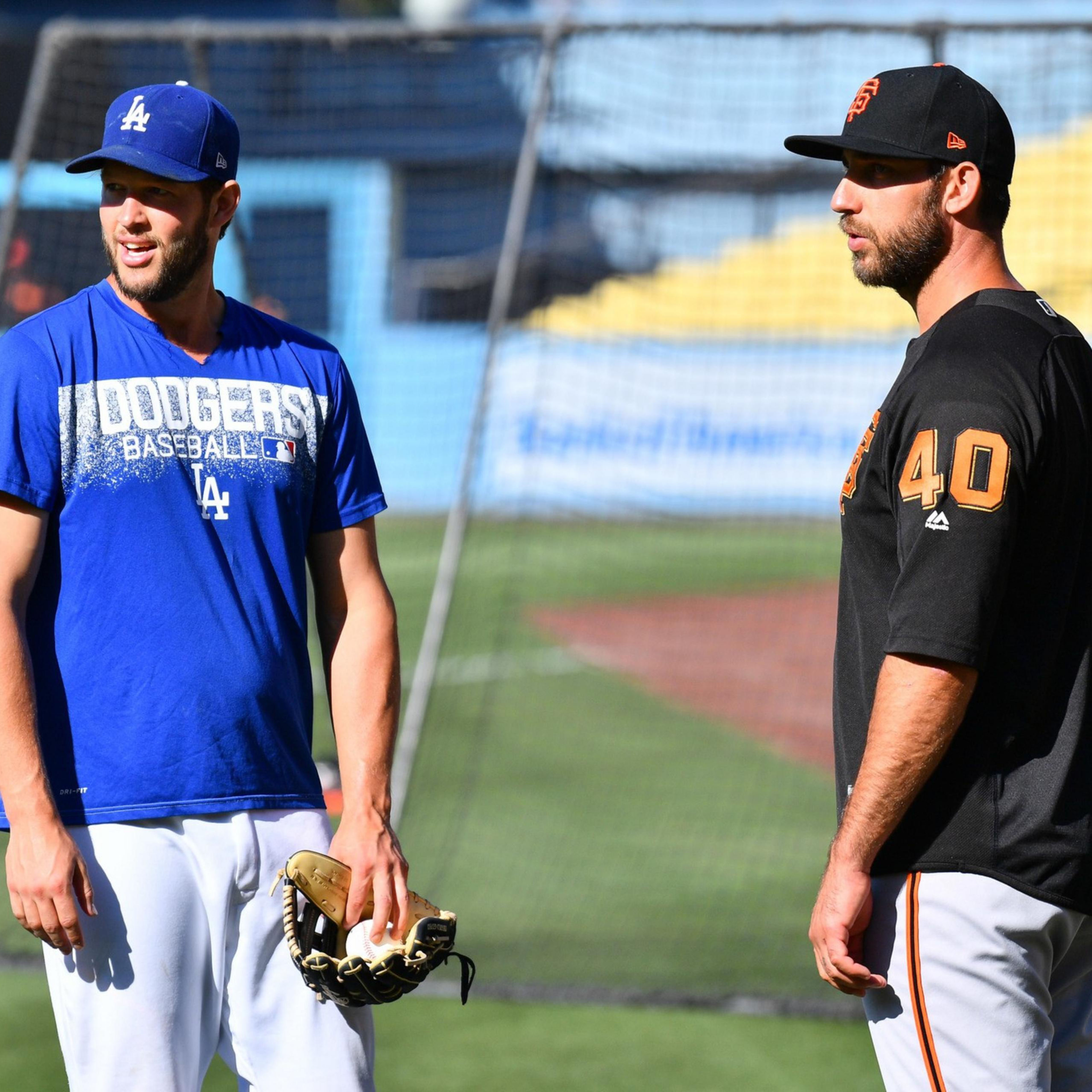 Two baseball players stand on a field, one in a blue Dodgers shirt and cap holding a glove, the other in a black Giants jersey and cap.