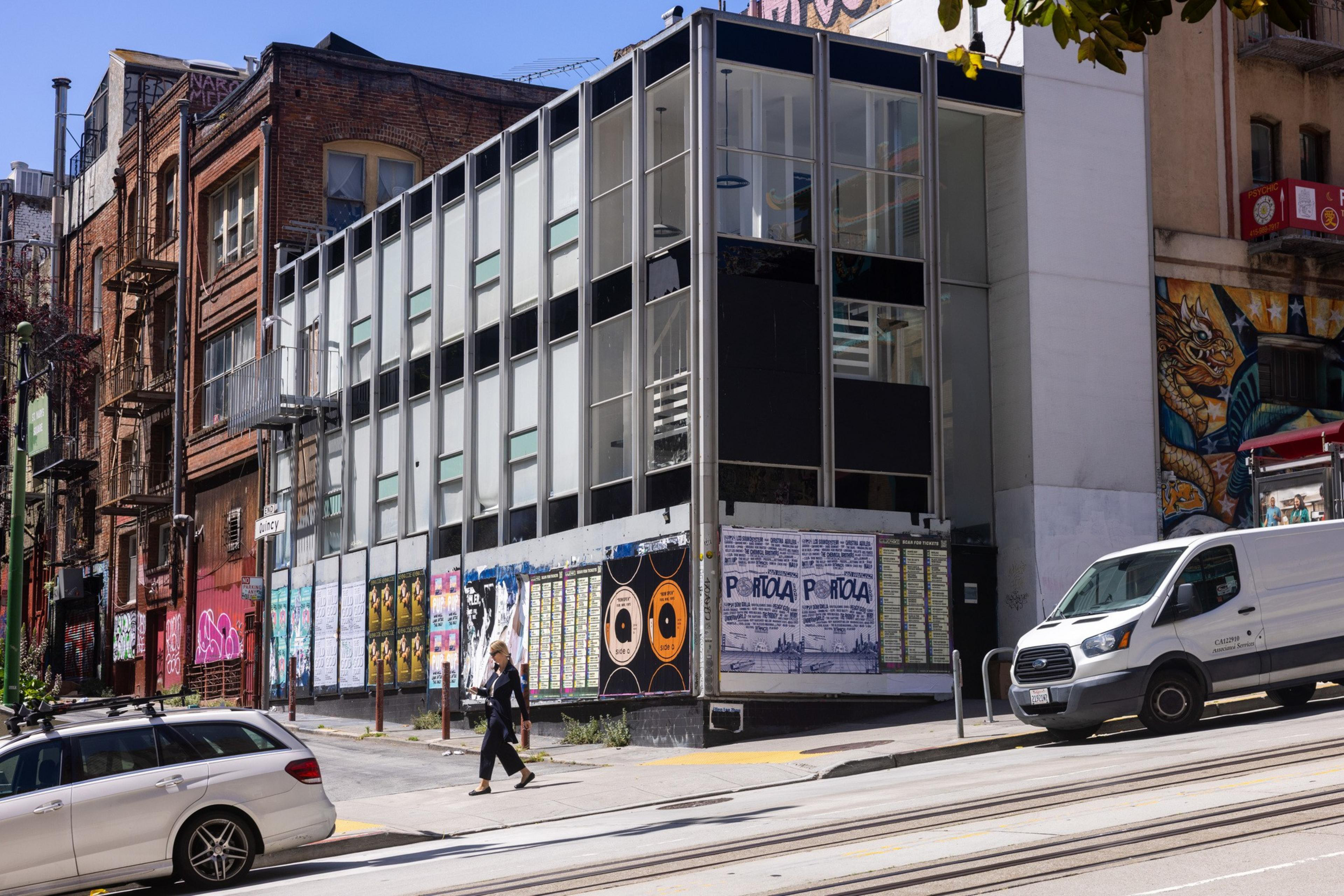 A modern glass building on a street corner has posters on its walls. Nearby, brick buildings stand. A woman walks across the street, with parked vehicles nearby.