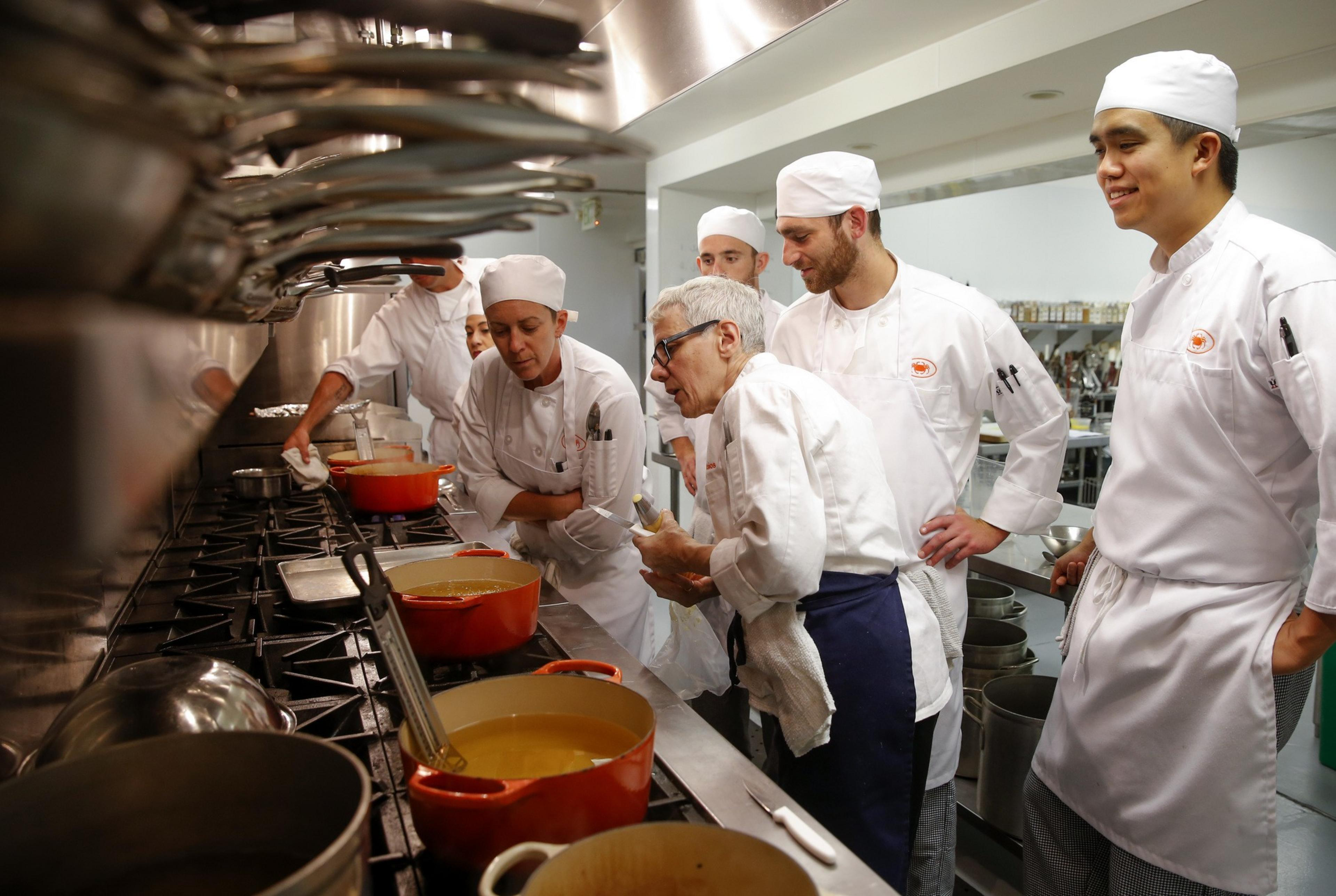 A group of chefs in white uniforms gather around a stove, focusing on cooking pots filled with liquid. They seem to be discussing or learning a technique.