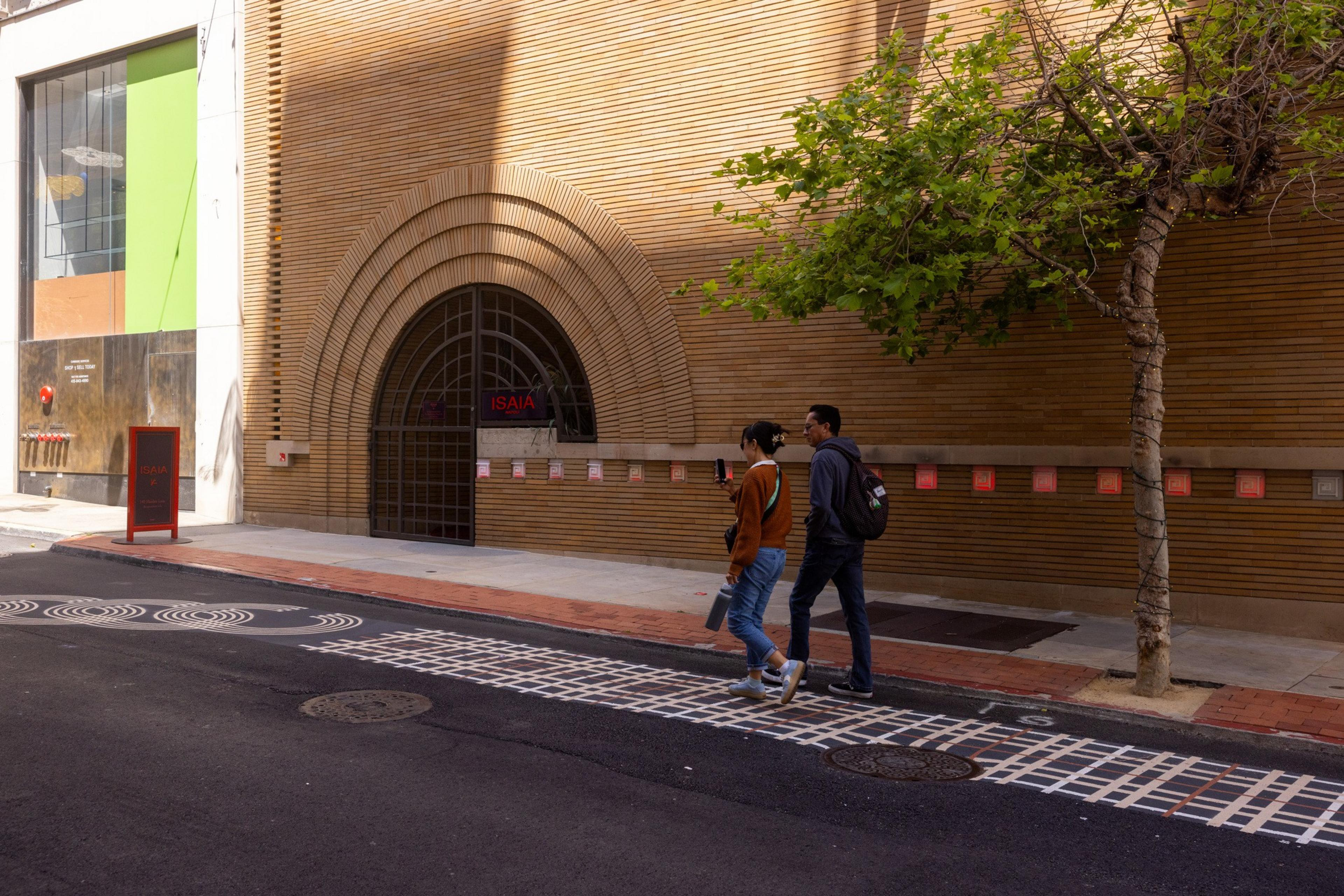 Two people walk on a sidewalk past a building with a large arched doorway labeled &quot;ISAIA.&quot; There's a tree and a green and orange window display nearby.