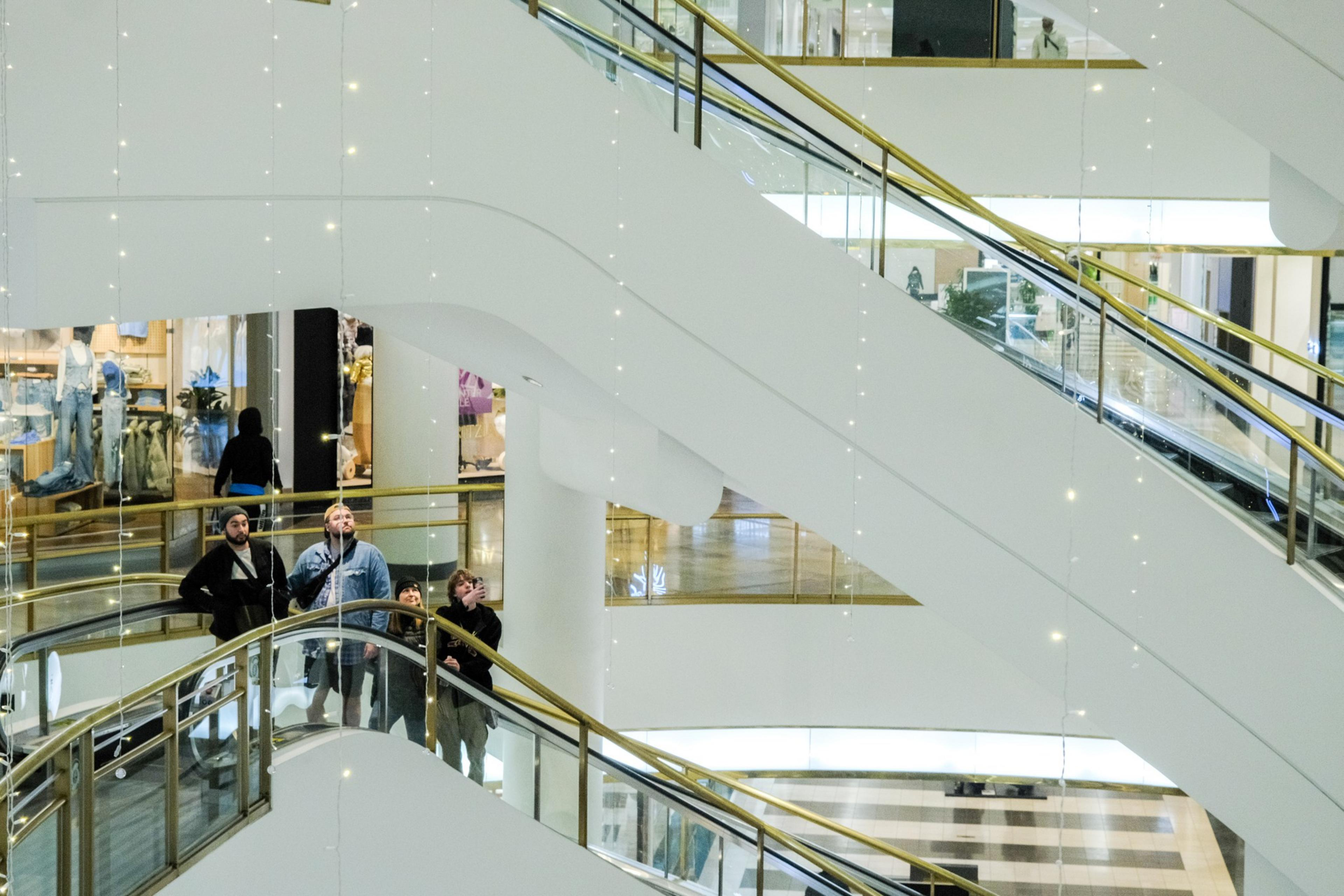 A multi-level shopping mall features several escalators and decorative lights. People stand on one escalator, looking upward, while shops are visible in the background.