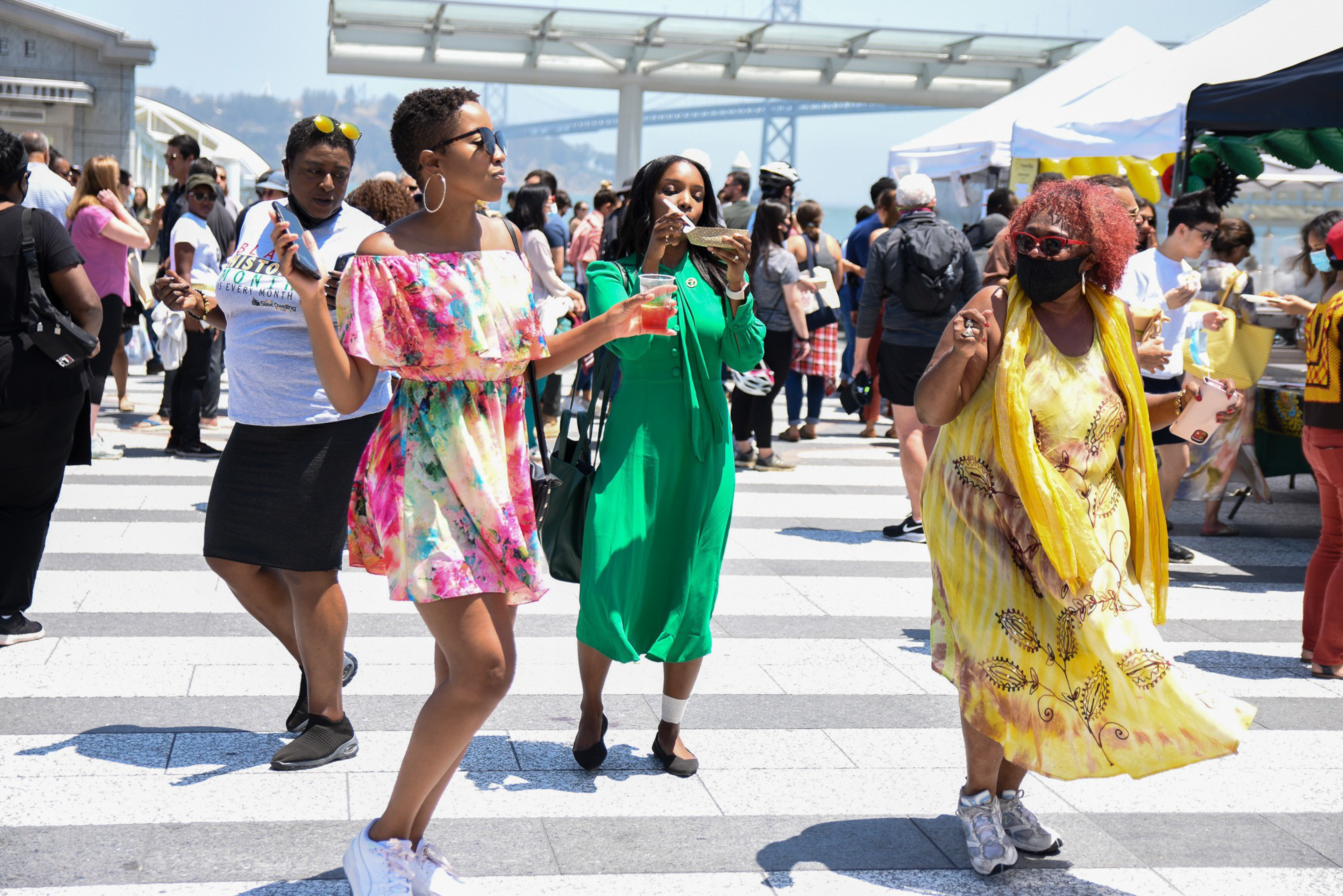 Four women wearing colorful dresses are enjoying themselves outdoors at a sunny event with many other people and tents in the background.