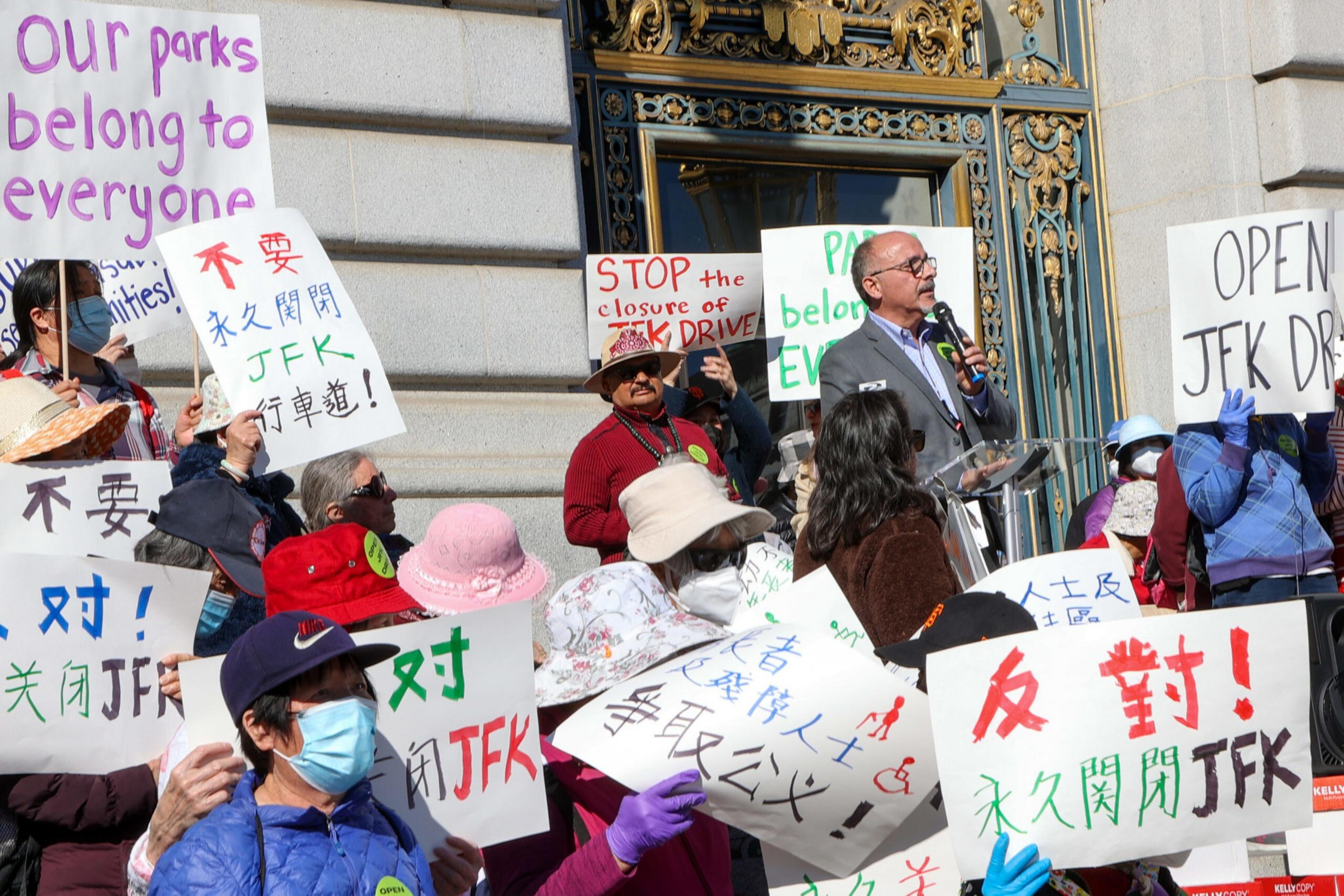 People gathered outside a building hold signs opposing the permanent closure of JFK Drive, with messages in English and Chinese, while a man speaks at a podium.