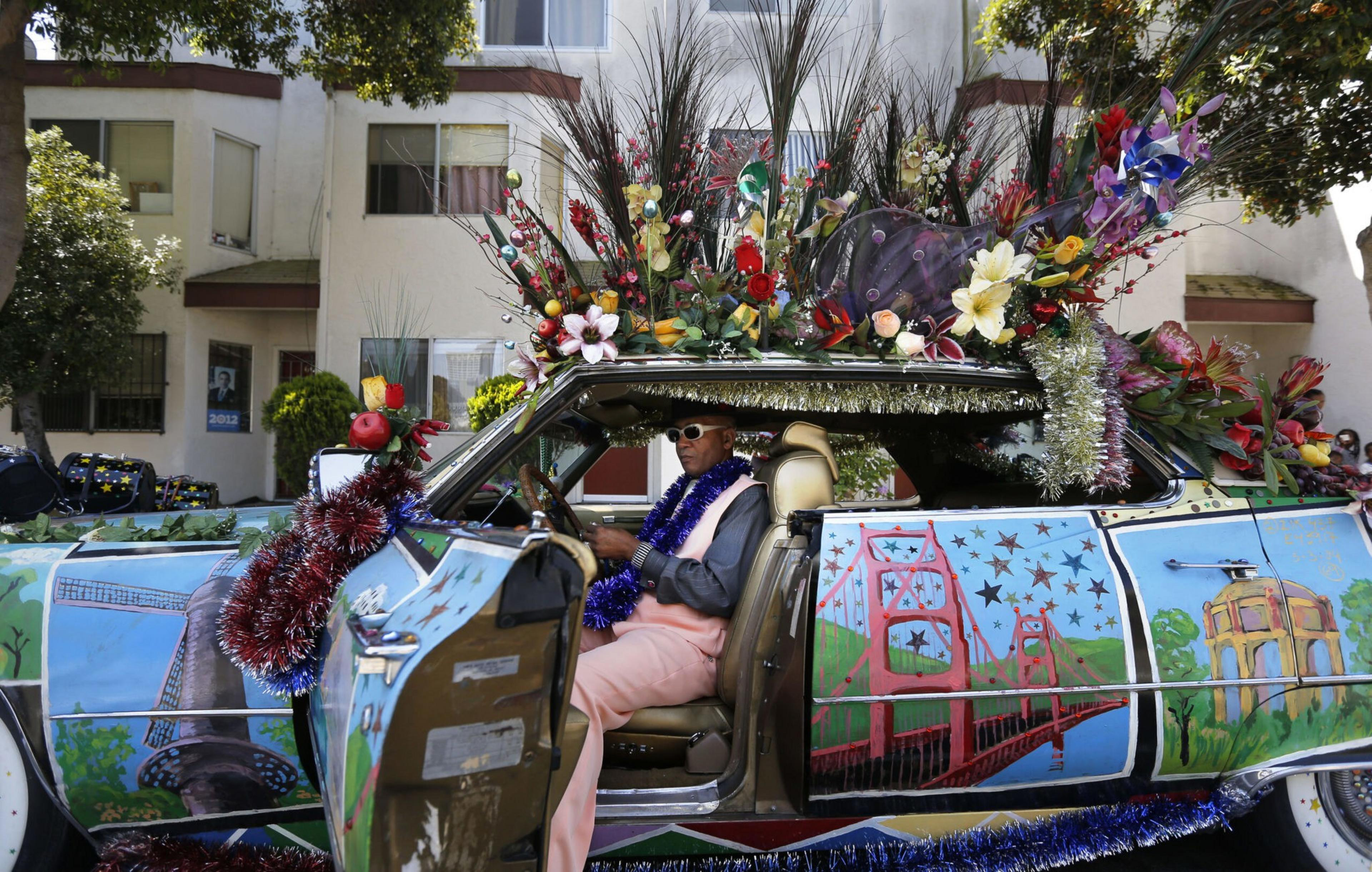 A man wearing sunglasses and bright clothes sits in a car covered with painted cityscapes and topped with colorful flowers and decorative branches.
