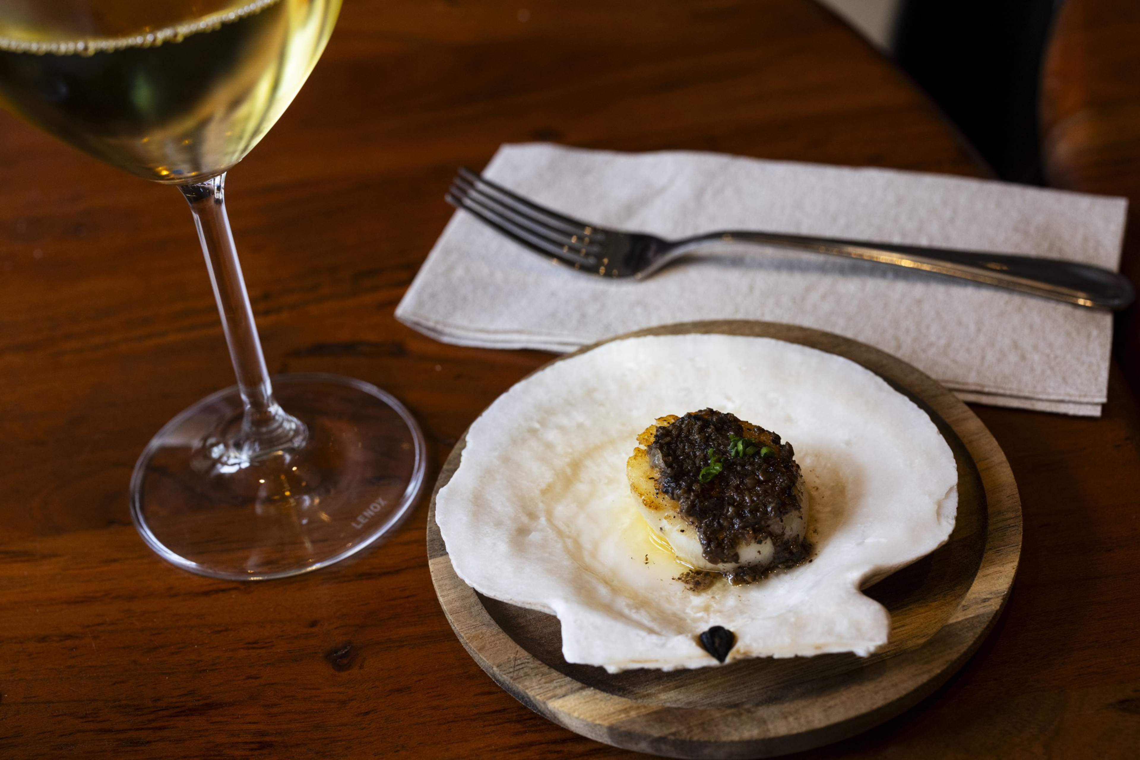 A seared scallop topped with black truffle sits on a white shell on a wooden plate, with a fork on a napkin and a glass of white wine nearby.