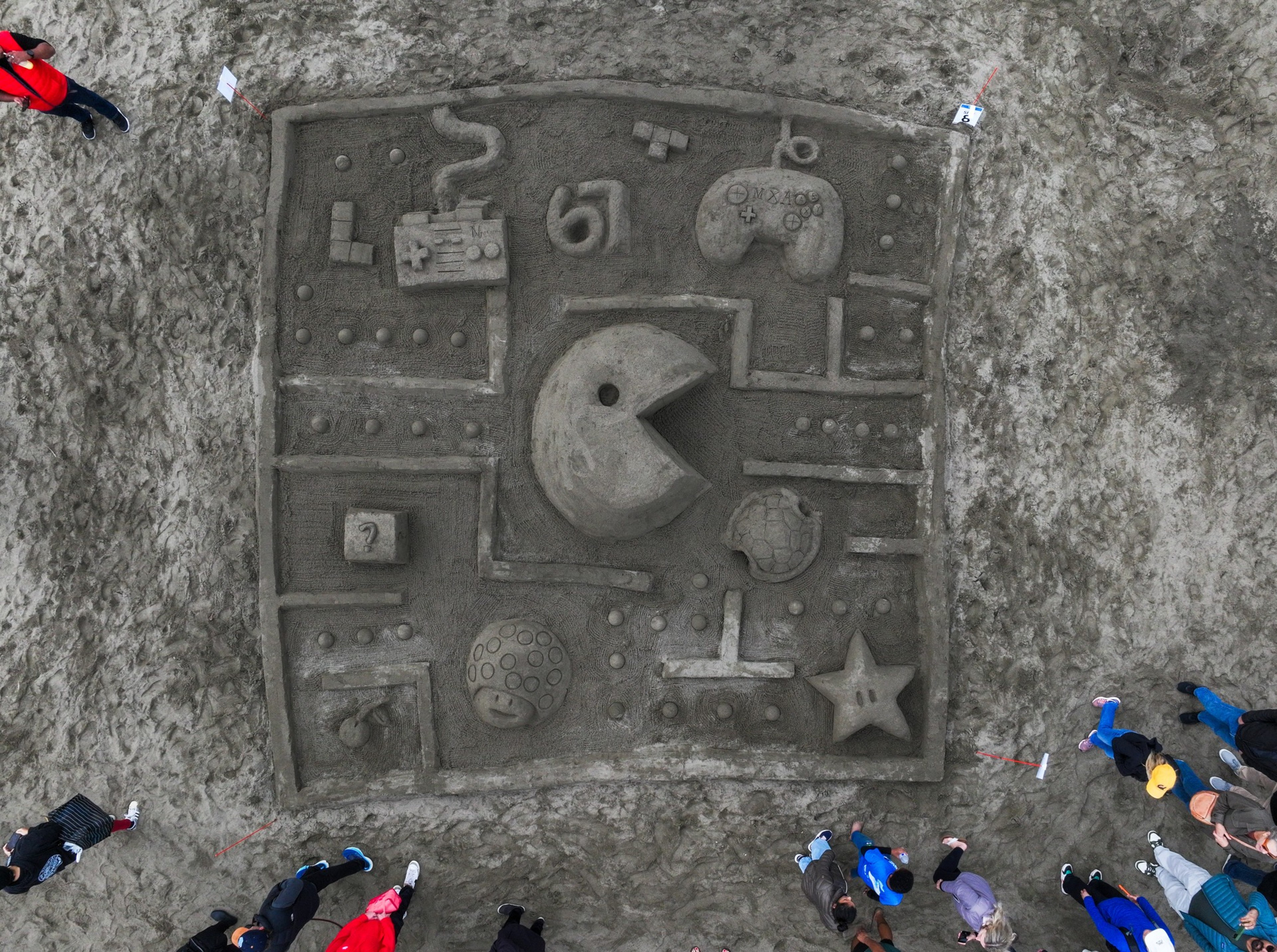 A large sand sculpture of a Pac-Man maze features Pac-Man, ghosts, a game controller, a question block, and other gaming icons, surrounded by spectators.