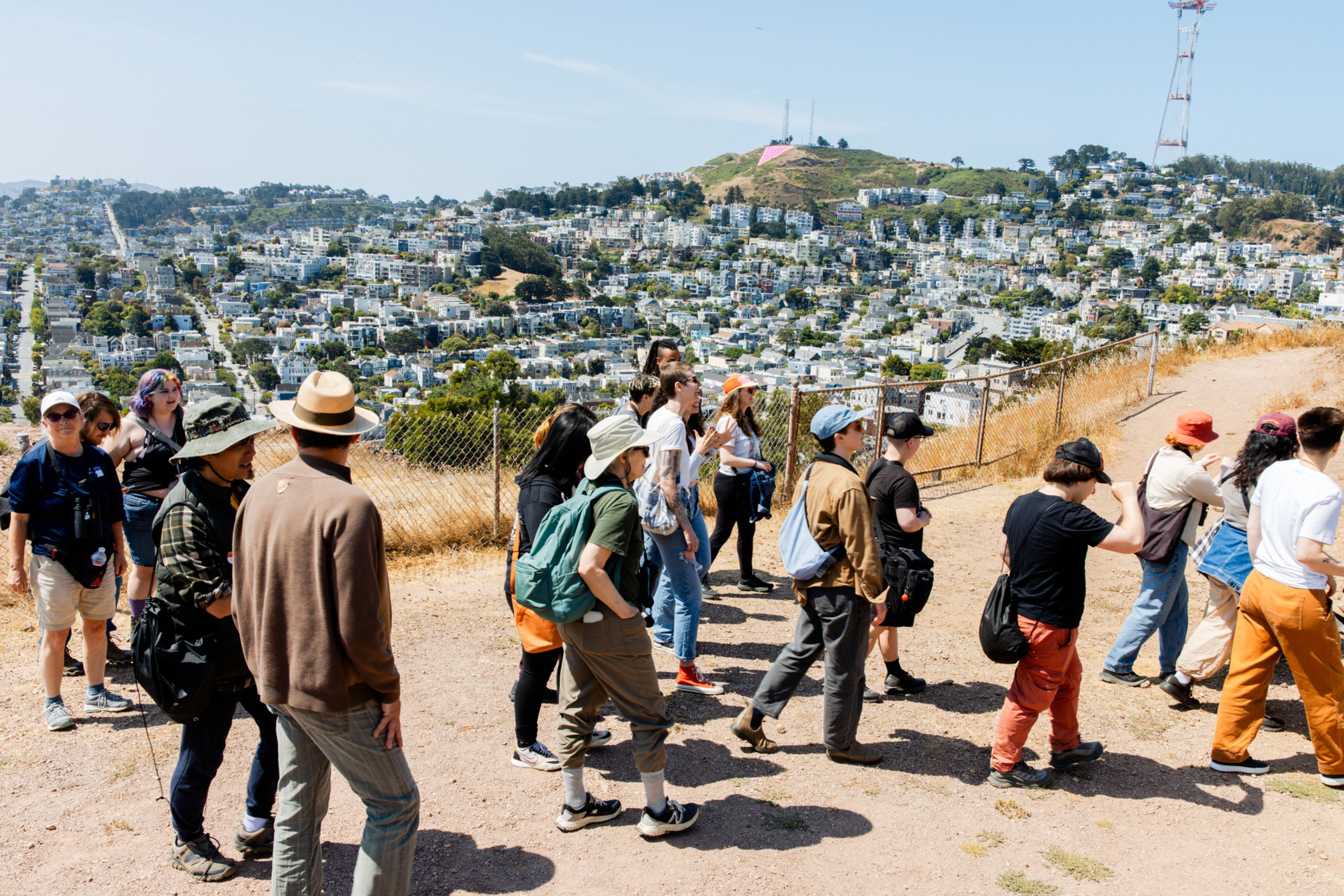 A group of people stands on a hill overlooking a city with densely packed buildings. They're near a fence, with a sunny sky above and communication towers visible.