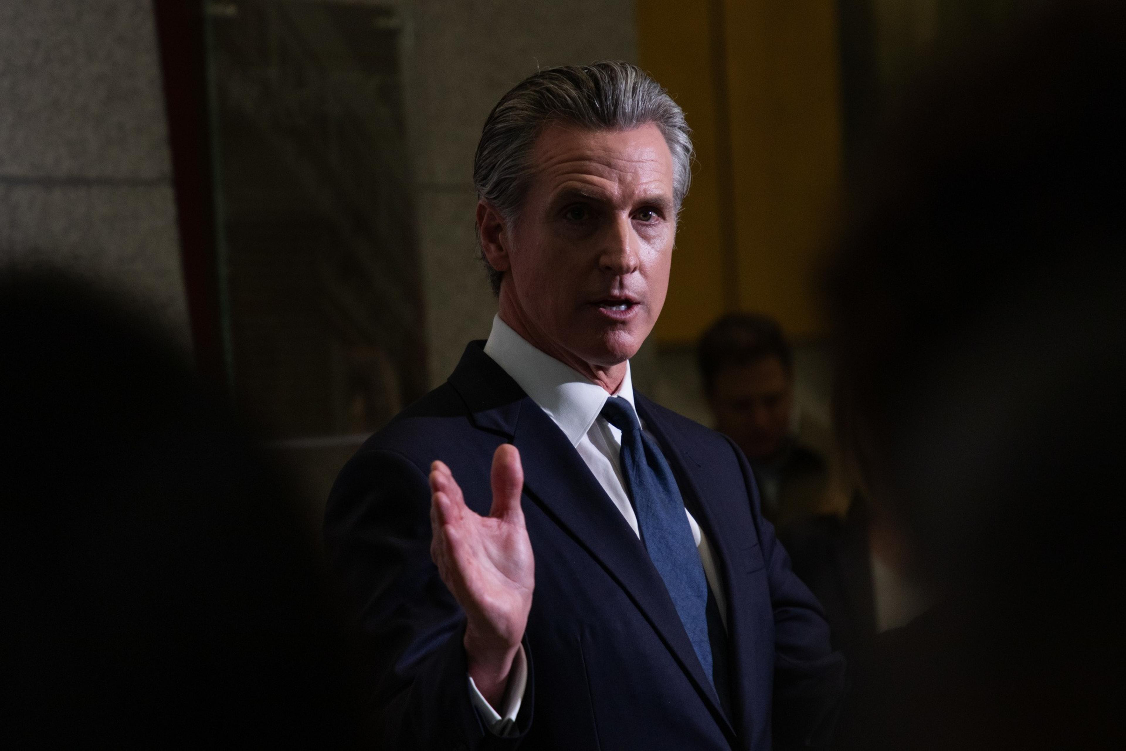 A man in a suit is speaking with an expressive hand gesture. He has short, gray hair and is in a dimly lit indoor setting, suggesting a formal event or meeting.