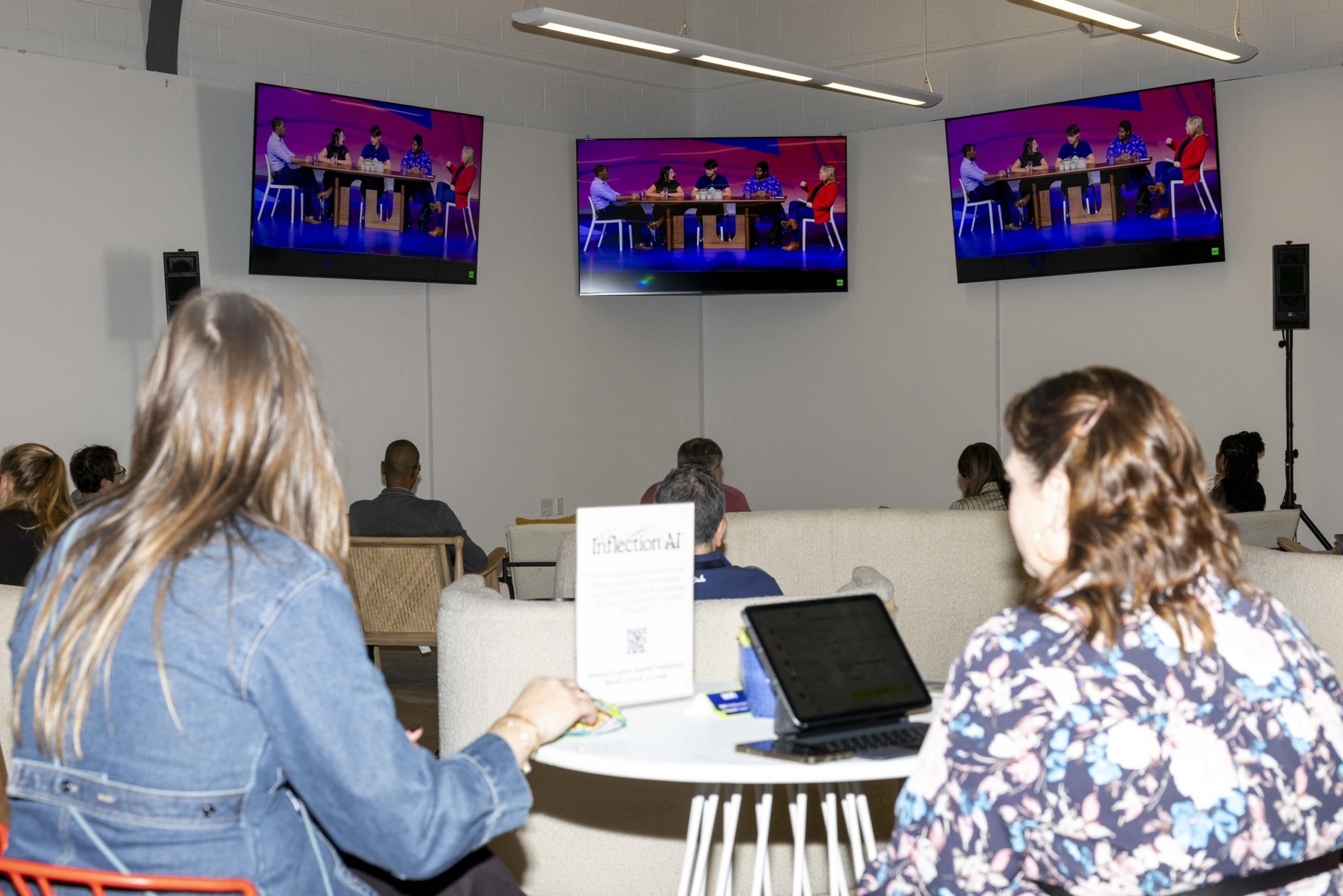 People are seated in a room watching a panel discussion on three large screens. A &quot;Inflection AI&quot; sign is visible on a table with a laptop.