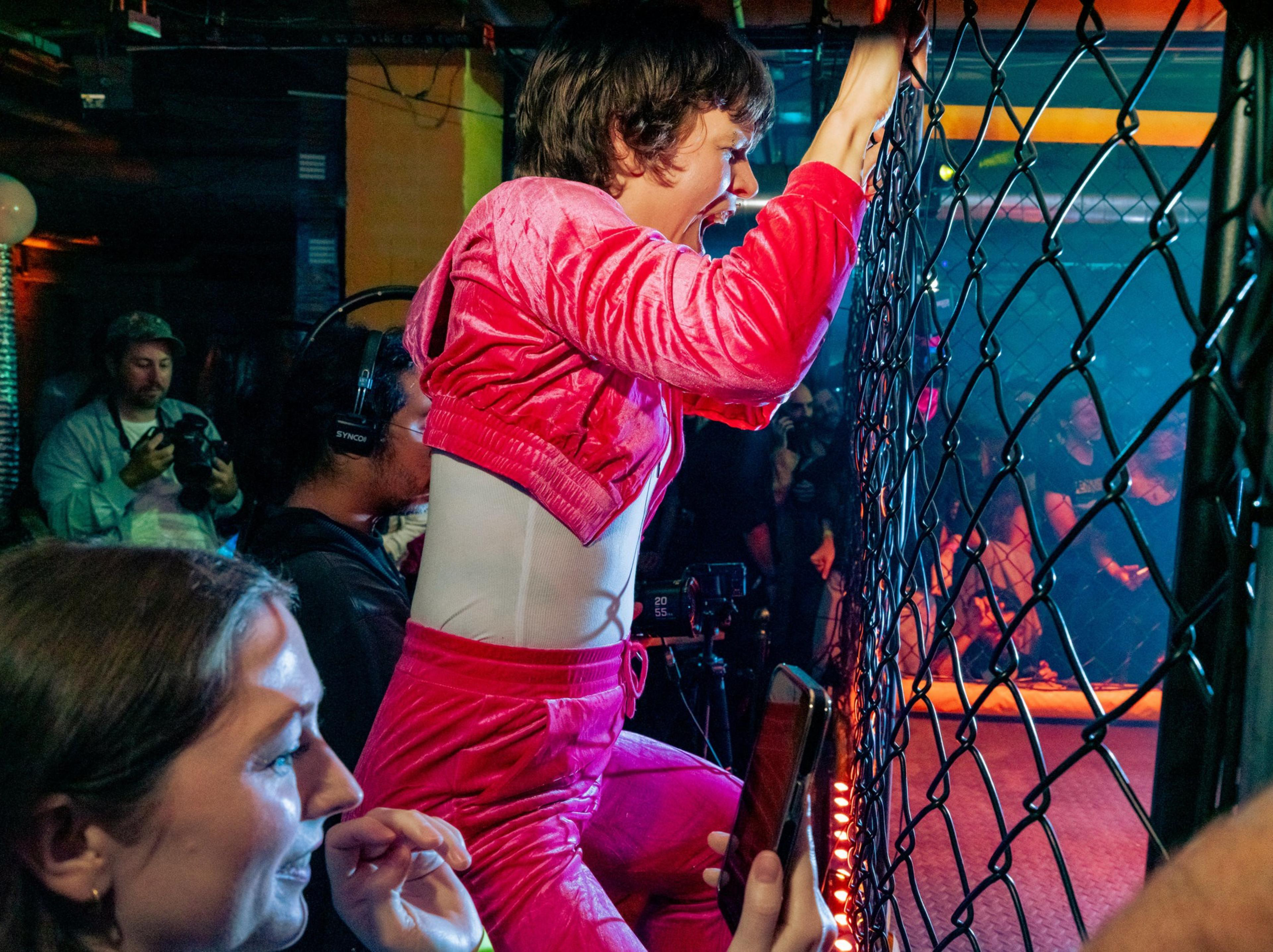 A person in a pink outfit grips a metal fence, shouting excitedly at an event, while others watch and record on their phones nearby.