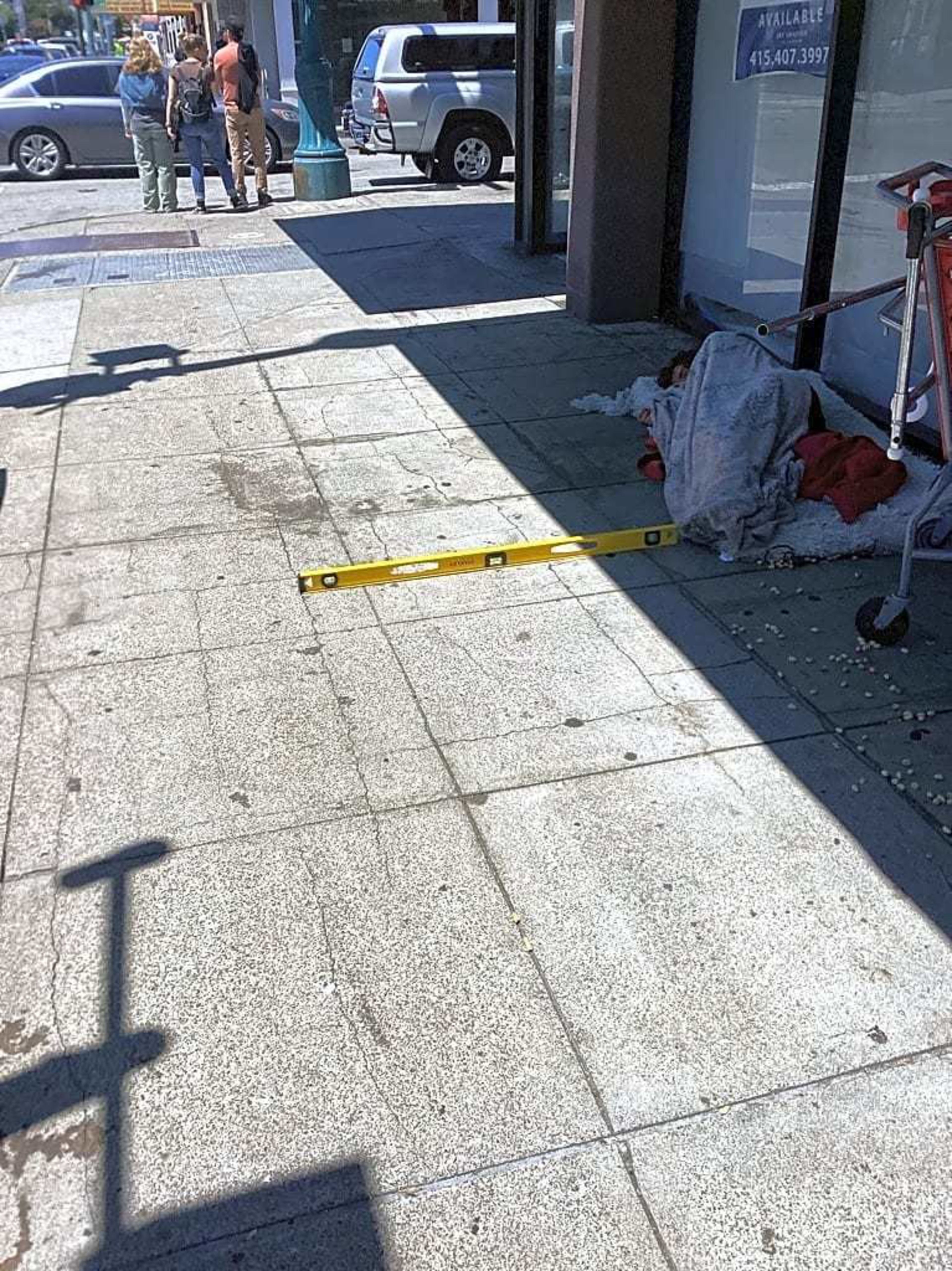On a sidewalk, a yellow level lies on the ground near an individual covered in a blanket, next to a shopping cart. Three people stand in the background by the street.