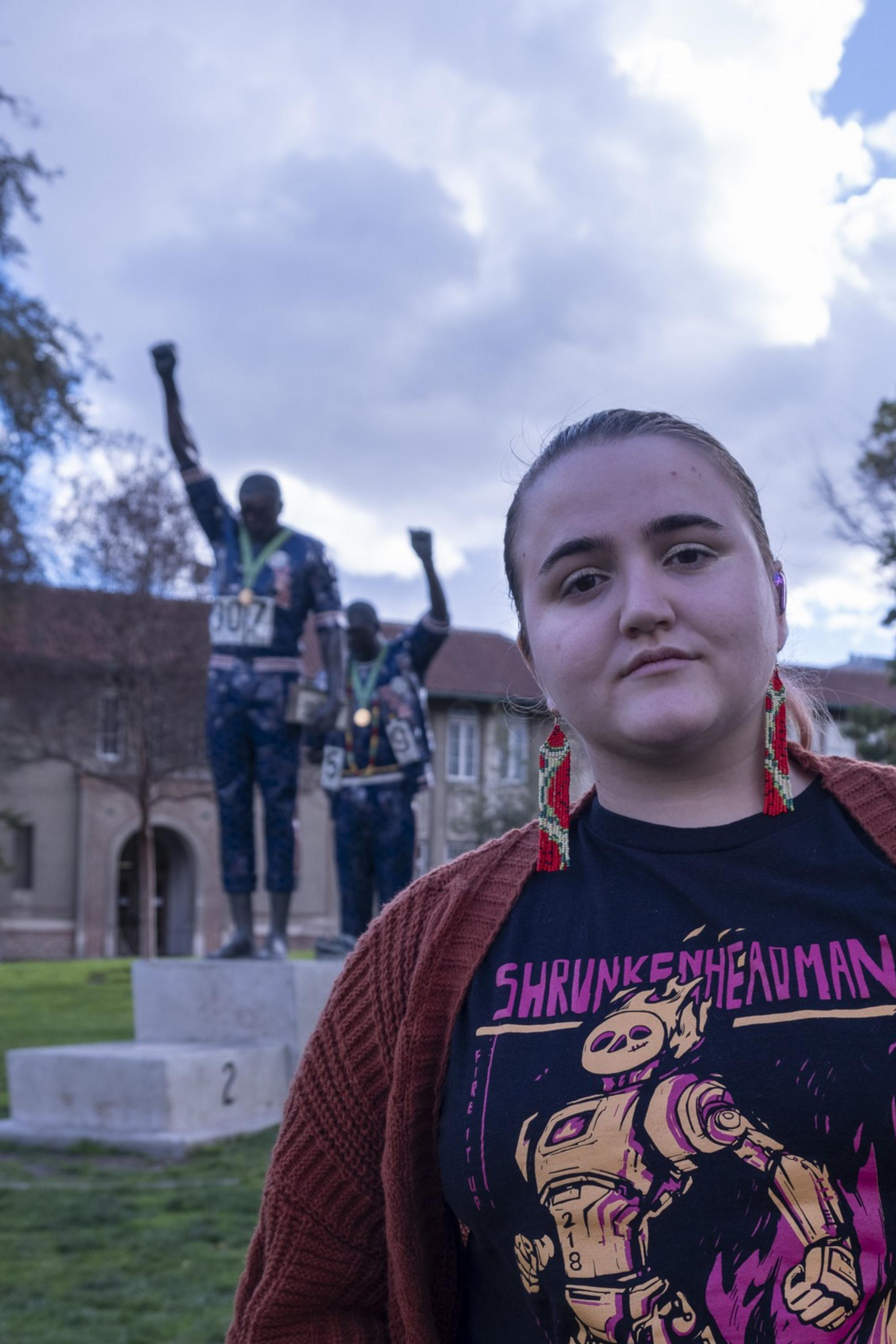 A person stands in the foreground wearing a graphic t-shirt and red cardigan. Behind them is a statue of two figures with raised fists, set against a cloudy sky.