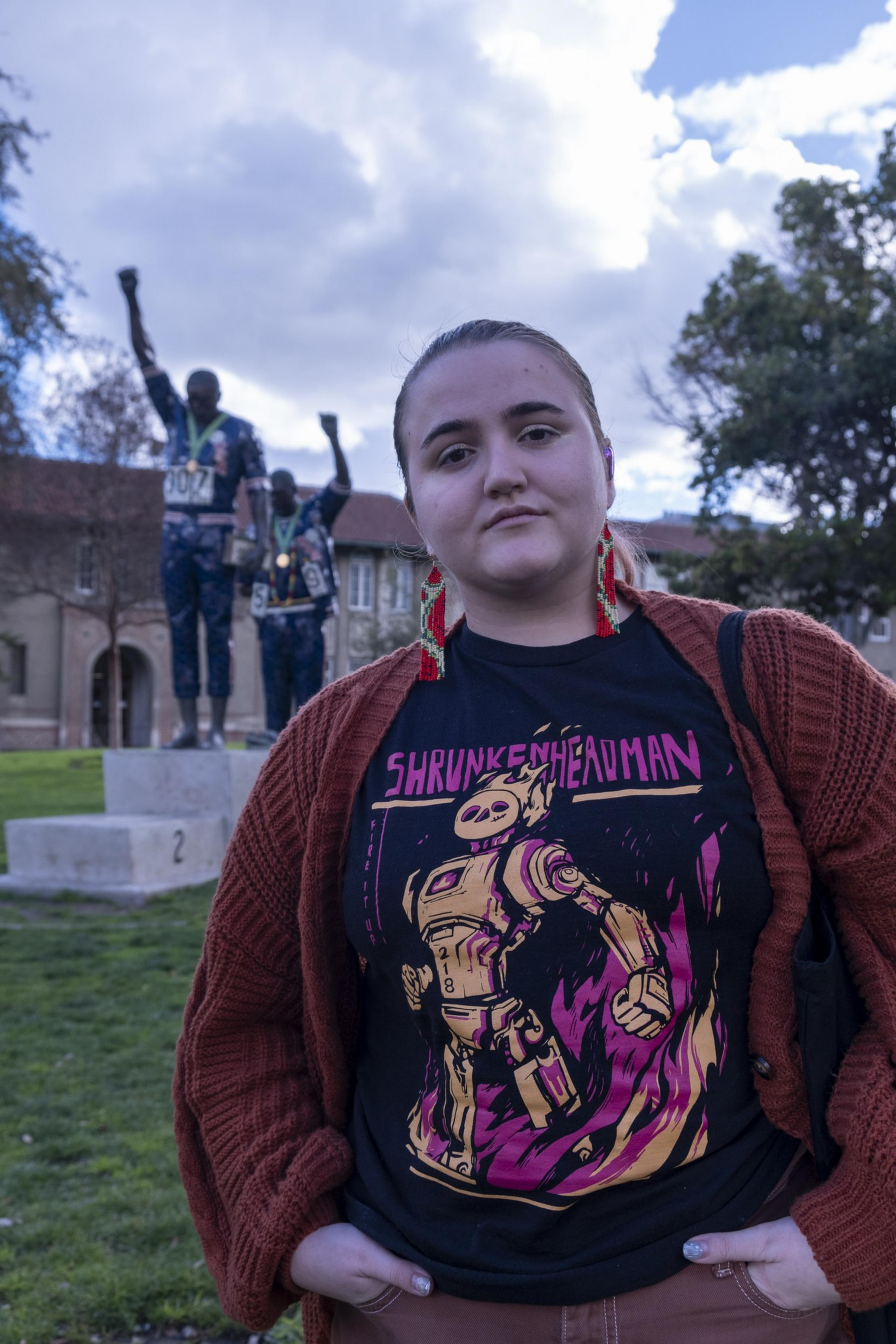 A person stands in the foreground wearing a graphic t-shirt and red cardigan. Behind them is a statue of two figures with raised fists, set against a cloudy sky.