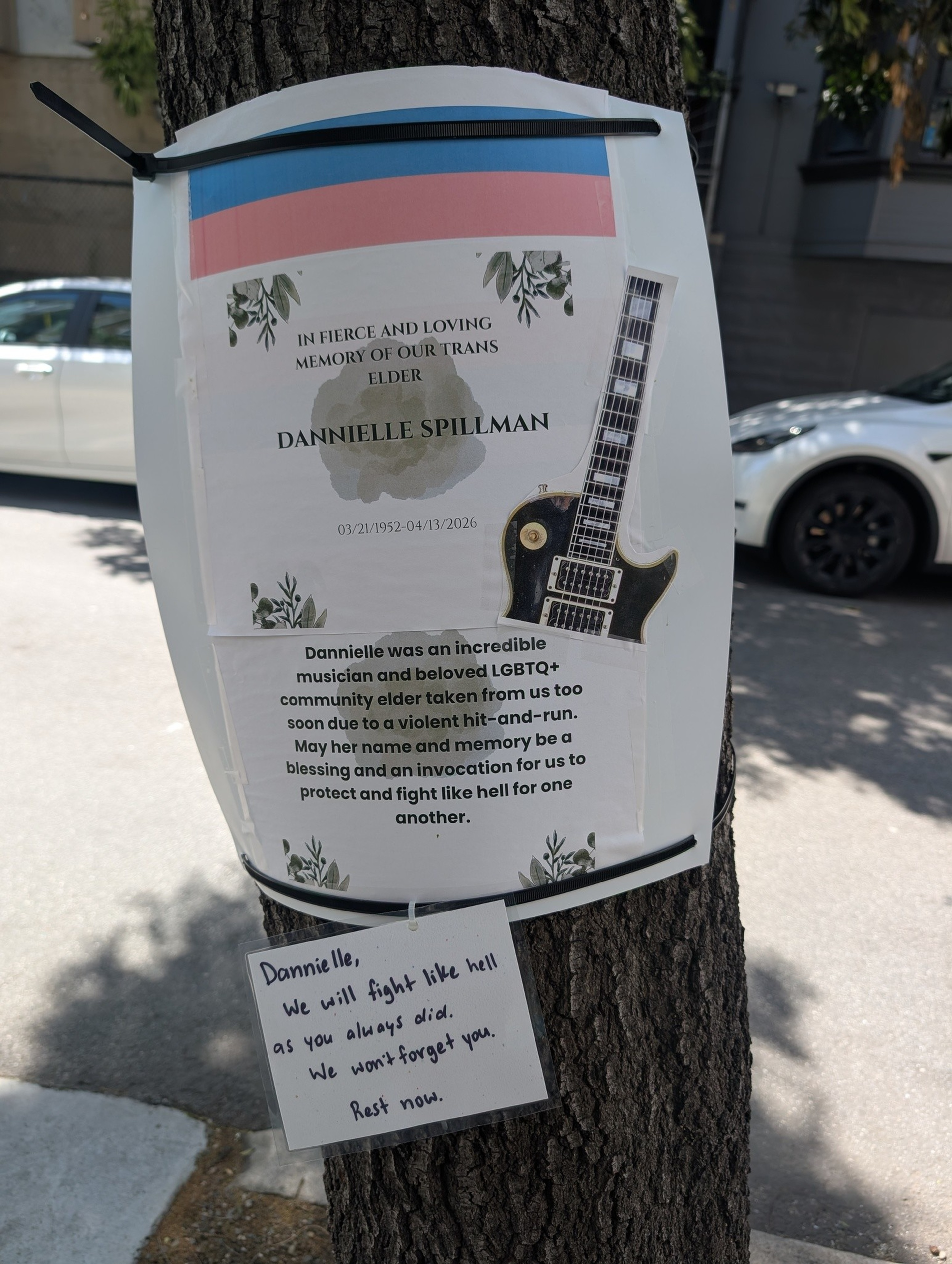 A memorial sign on a tree honors Danielle Spillman, a beloved trans elder and musician, killed in a hit-and-run, with a handwritten note pledging to continue the fight.