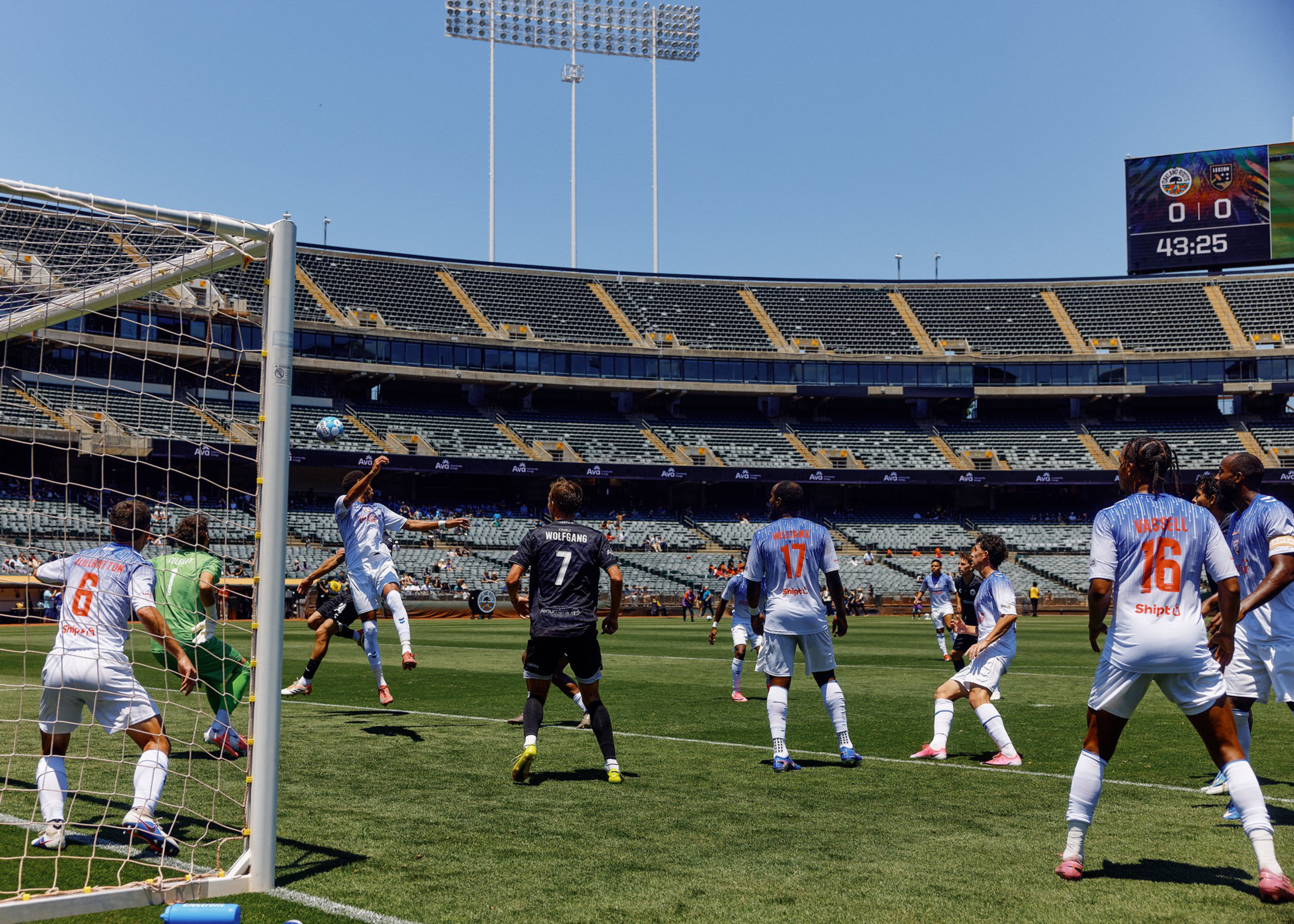 Soccer players from two teams compete near the goal, with one player jumping to head the ball as the goalkeeper prepares to defend.
