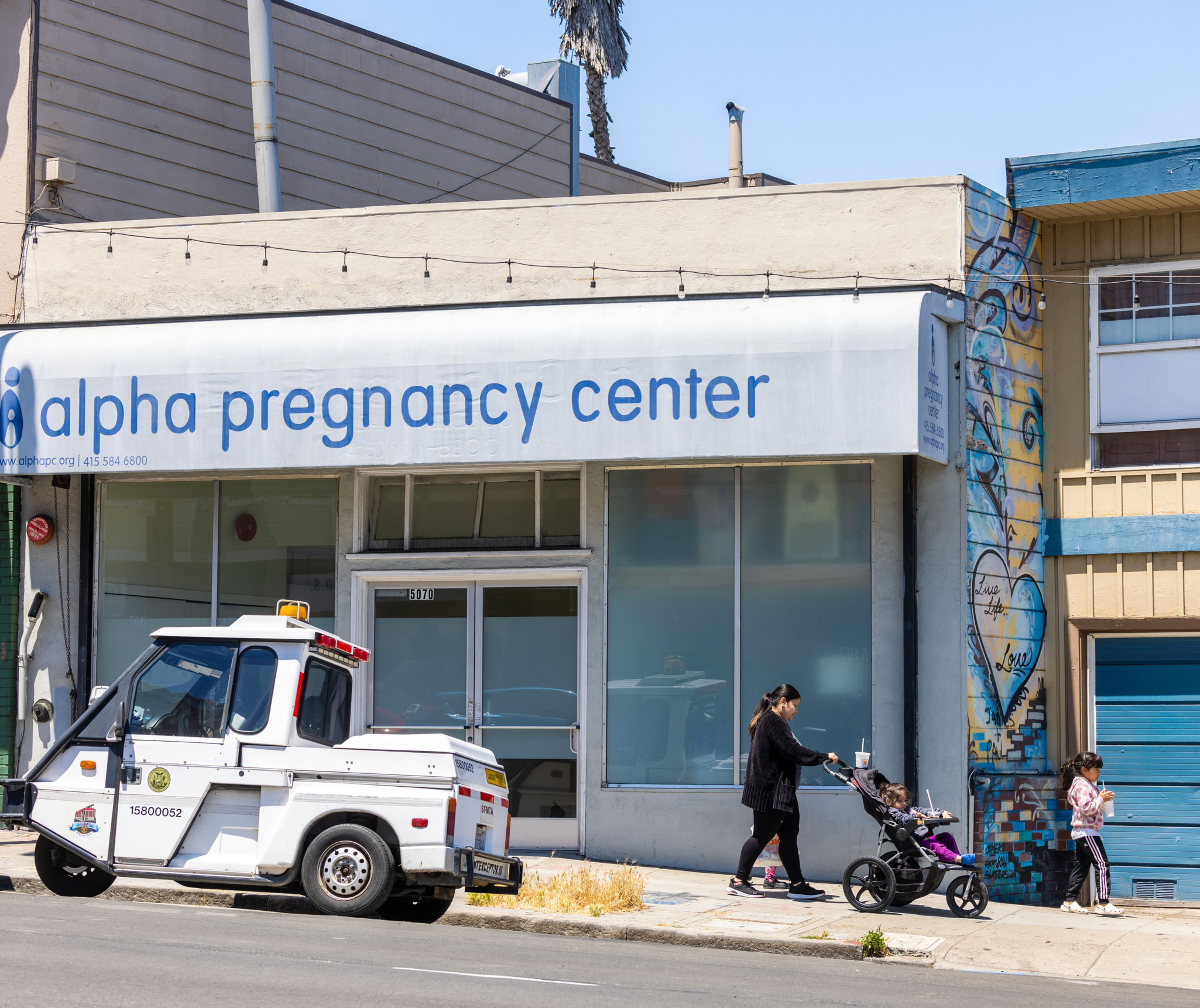 A woman pushes a stroller past a white building labeled &quot;alpha pregnancy center.&quot; A utility vehicle is parked in front, and two children stand near a mural on the wall.