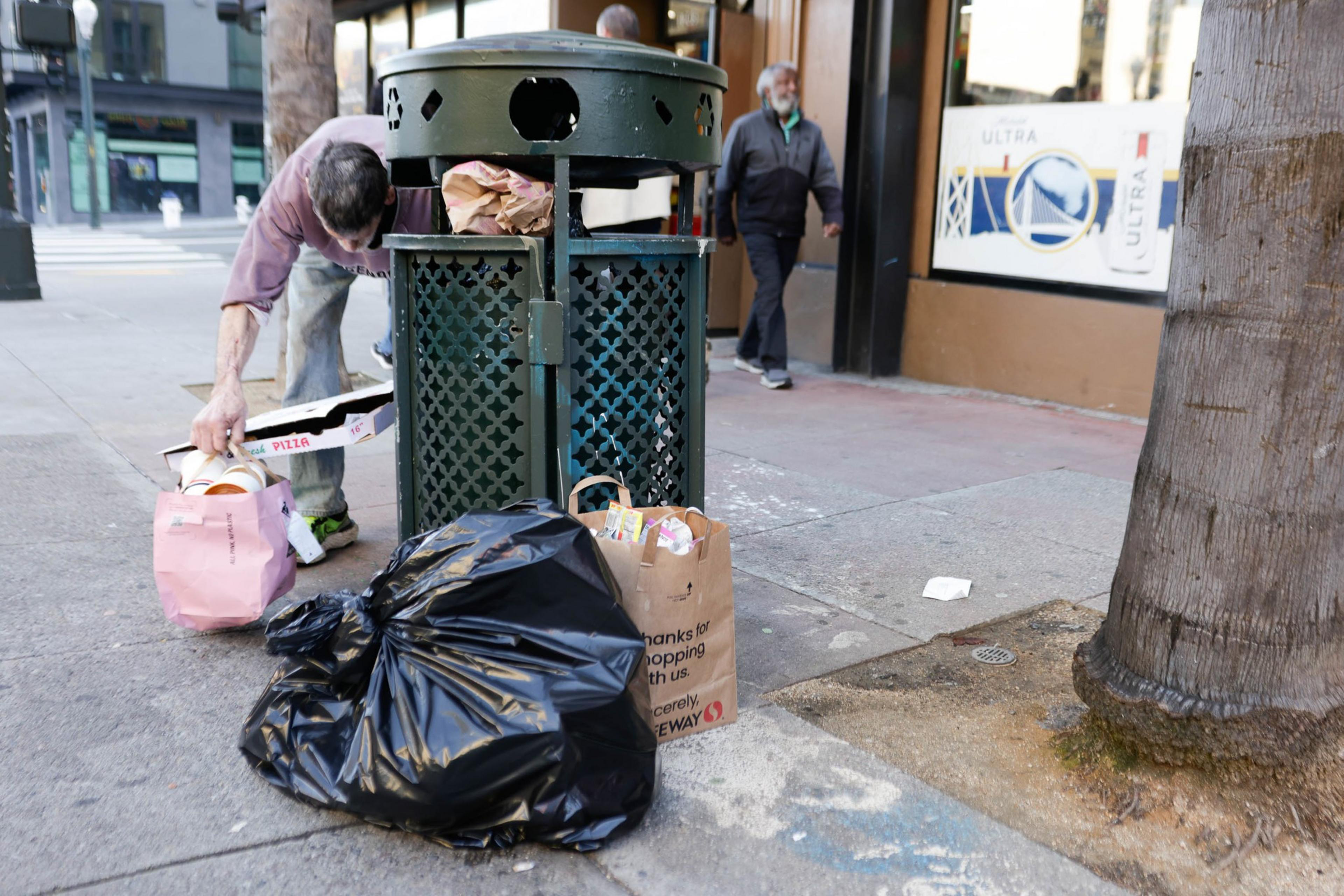A man places a pizza box and bag of trash next a public trash can.