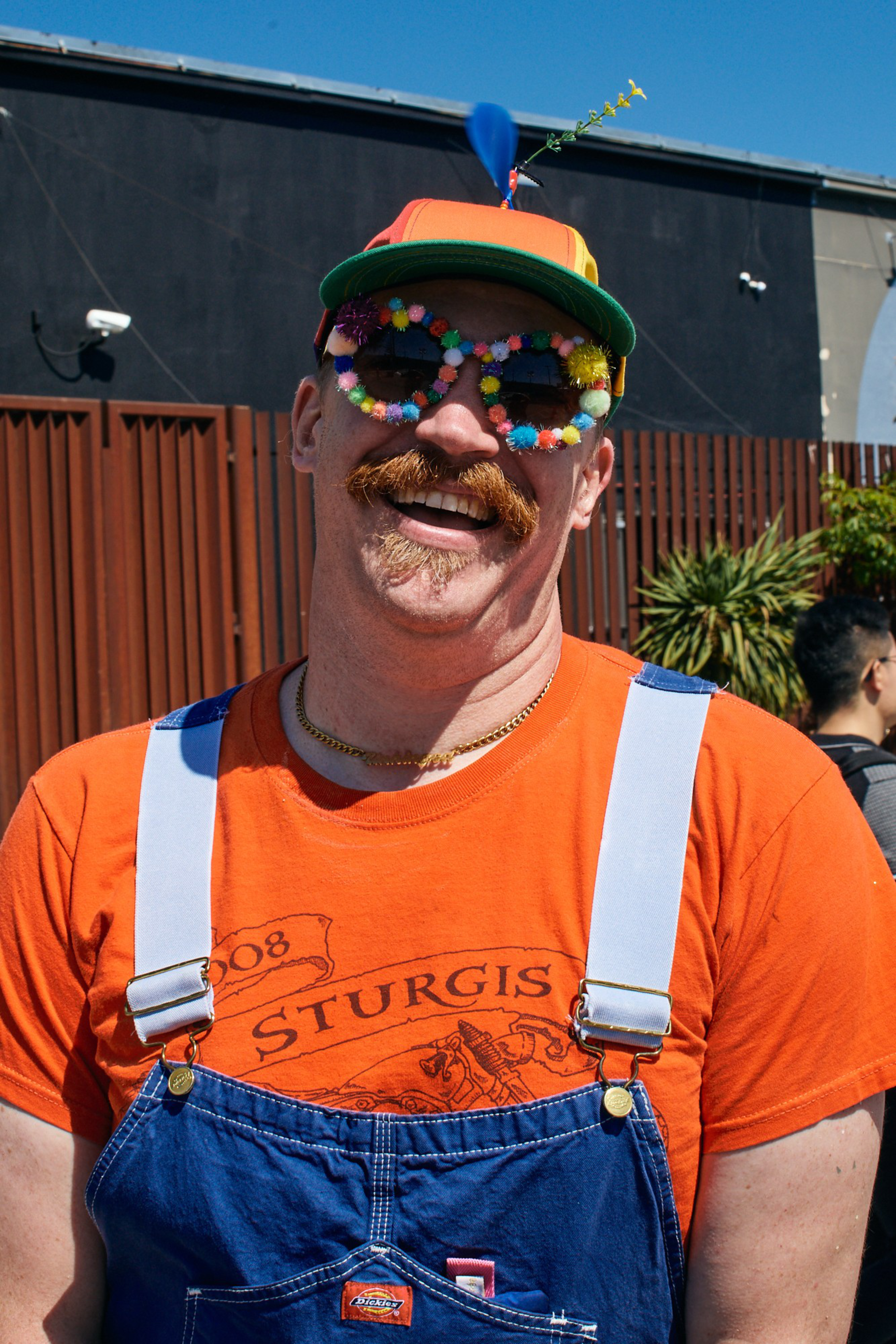 A man wearing colorful pom-pom sunglasses, an orange shirt, denim overalls, and a multicolored cap with a small dart stuck in it smiles outdoors.