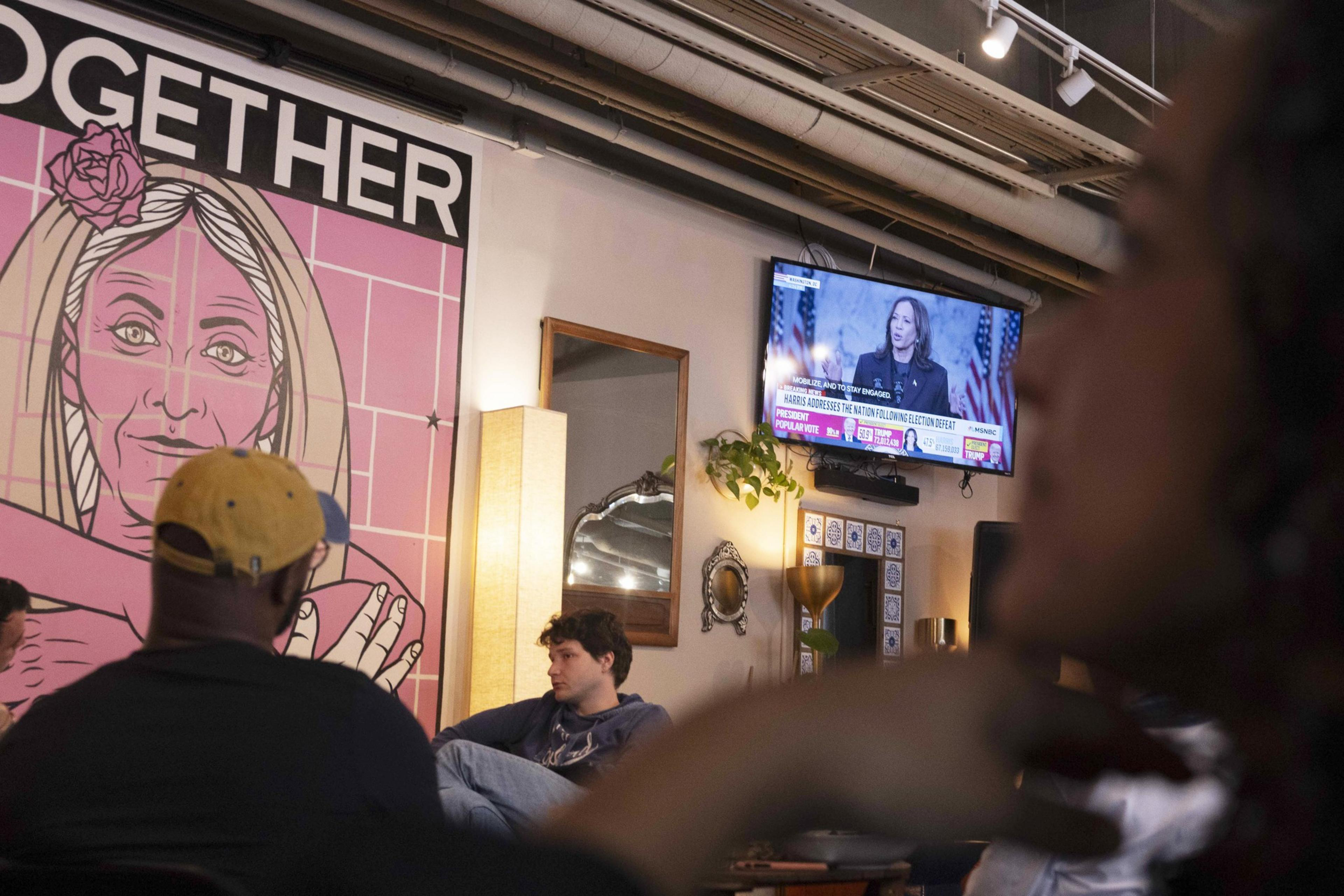 A casual setting with people seated, featuring a large pink mural of a person alongside a TV showing a news broadcast. The space is warmly lit.
