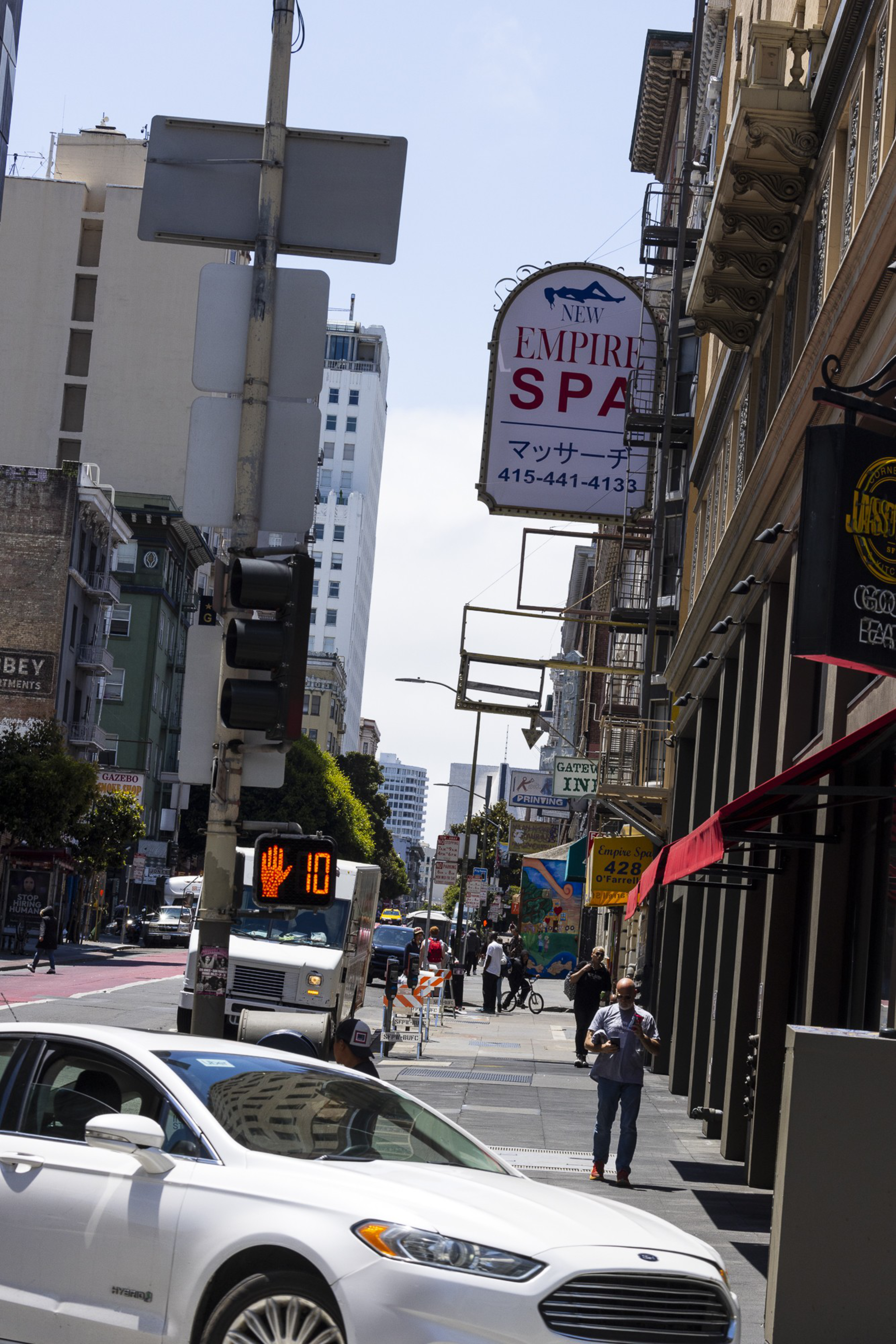 A city street scene shows people walking on sidewalks, a white car in the foreground, and various storefront signs. A traffic signal displays a countdown.