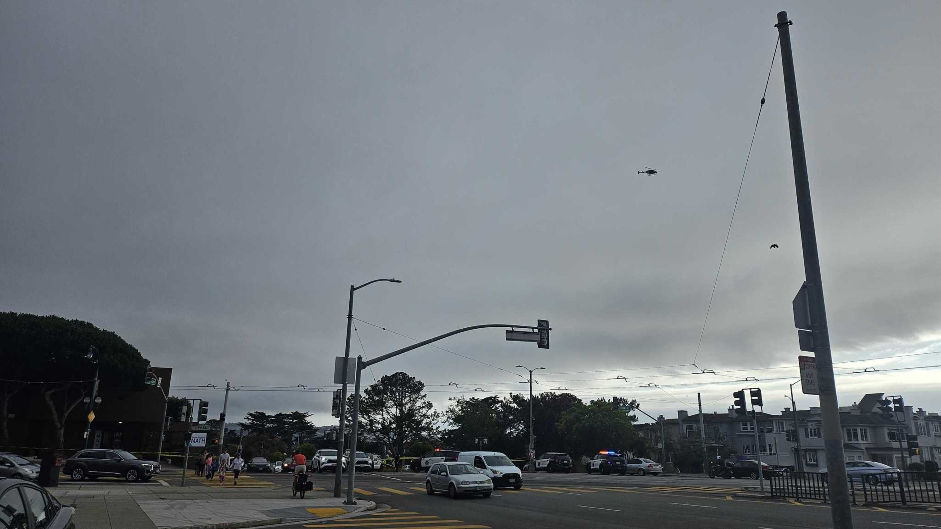 A gray, overcast sky looms over a busy urban street with cars, a motorcyclist, pedestrians, a traffic light, and a helicopter flying above.
