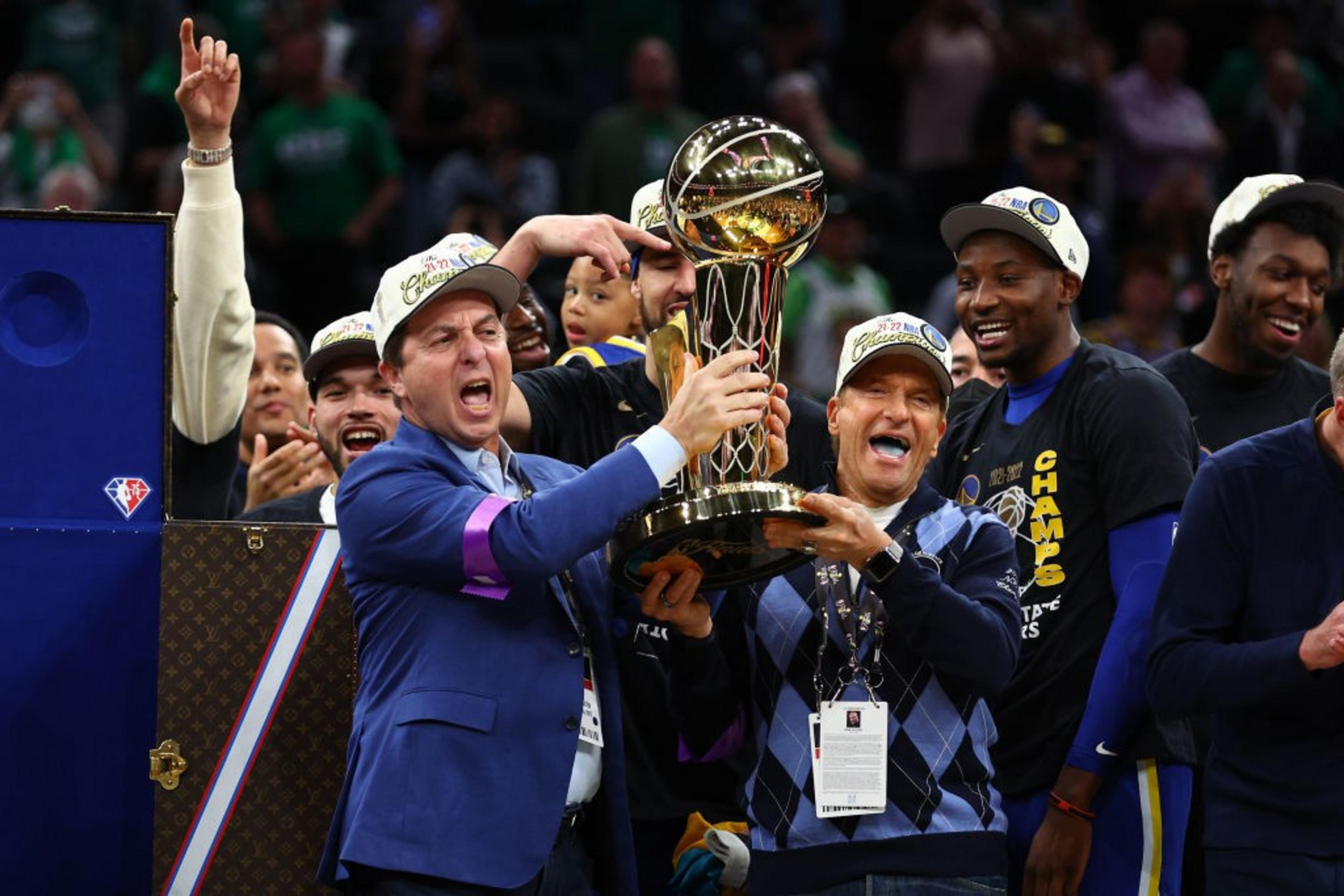 Two men jubilantly hold a large gold trophy, surrounded by a cheering group wearing championship hats and shirts, celebrating a significant sports victory.