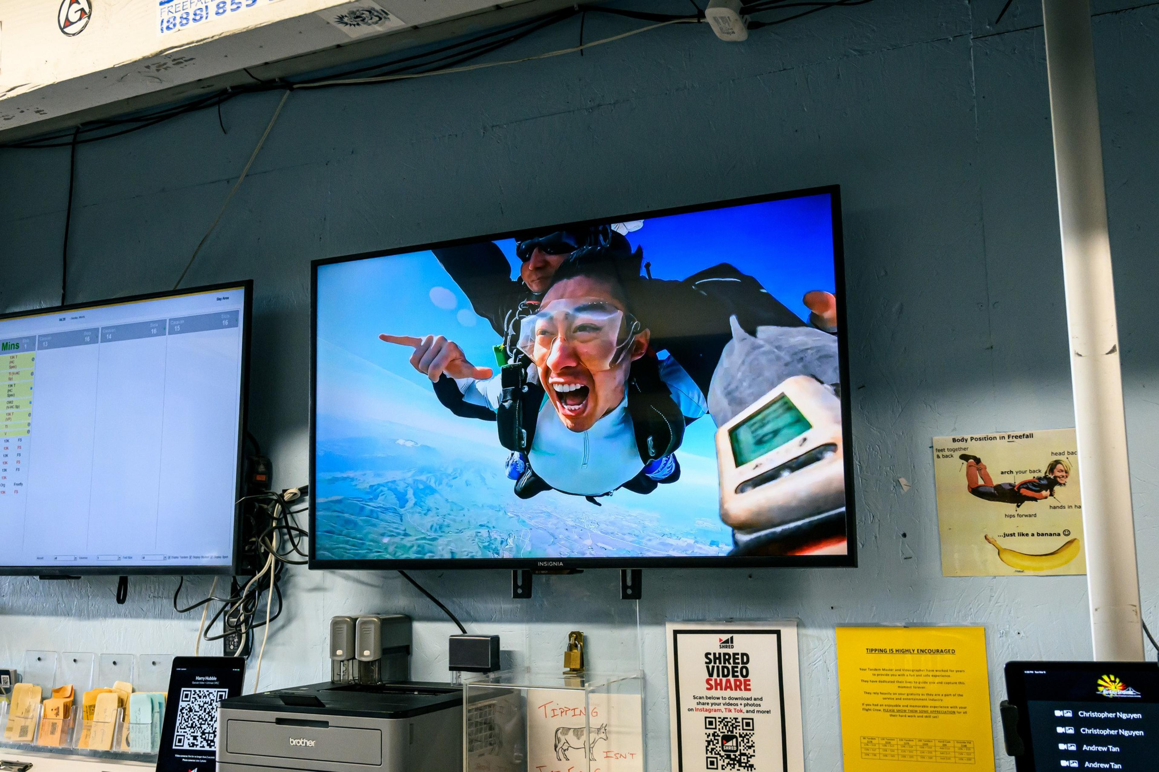 A TV screen displays a skydiving scene with a person excitedly free-falling tandem with an instructor, set against a blue sky and landscape background.