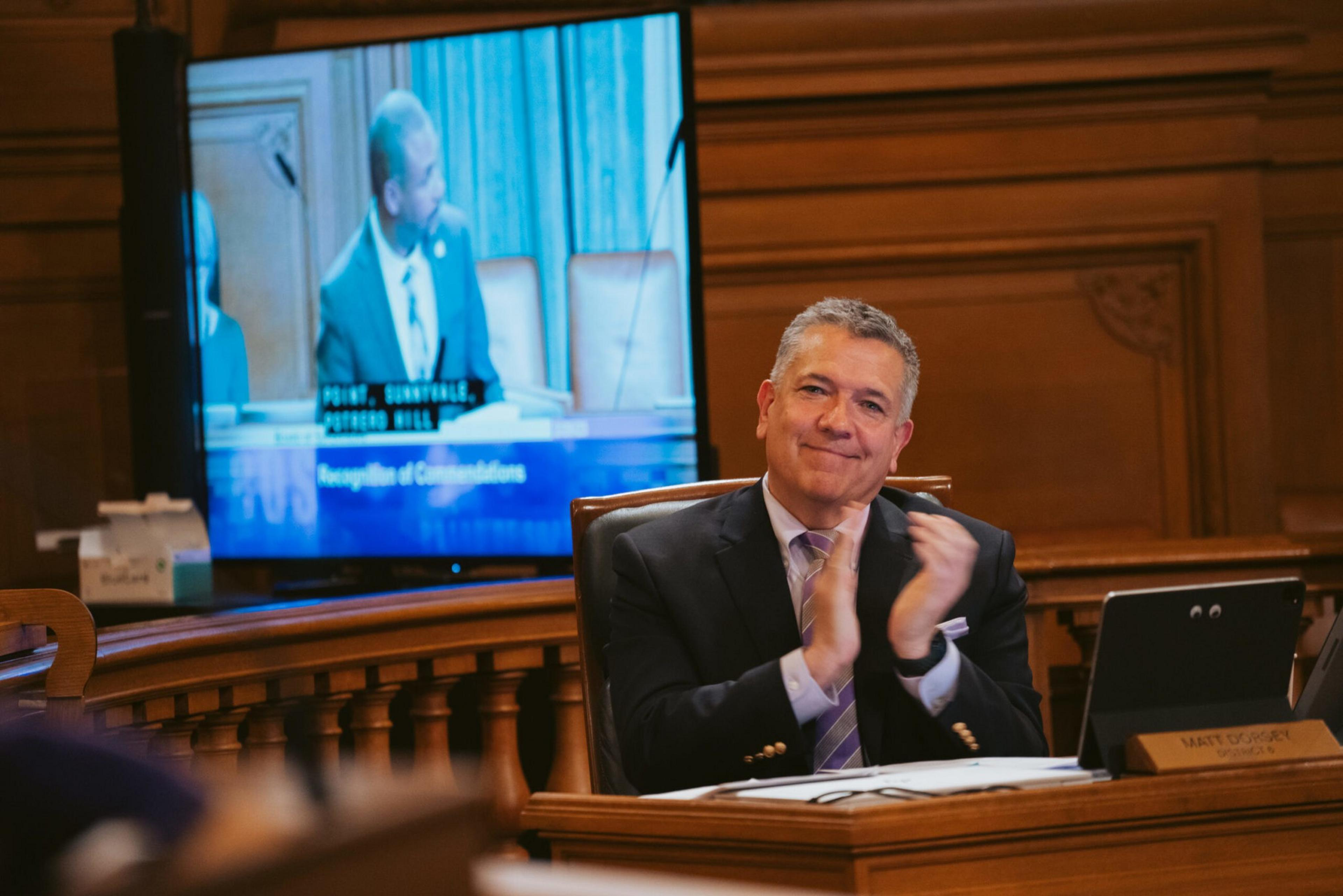 A man in a suit claps while sitting at a desk in a wood-paneled room, with a TV screen behind him showing another man speaking.