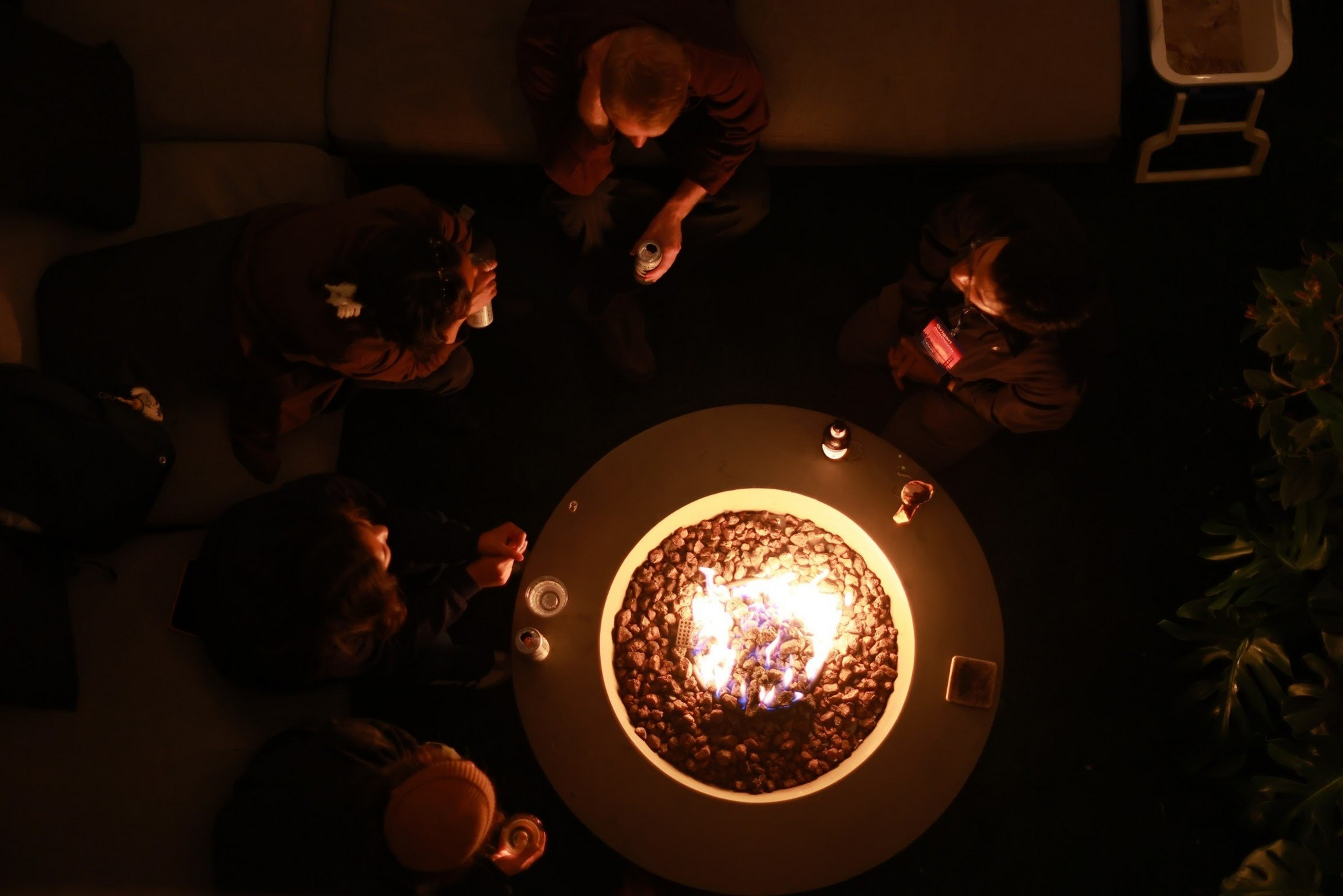 Five people sit around a round fire pit lit with bright flames, holding drinks, in a dimly lit outdoor setting with plants nearby.