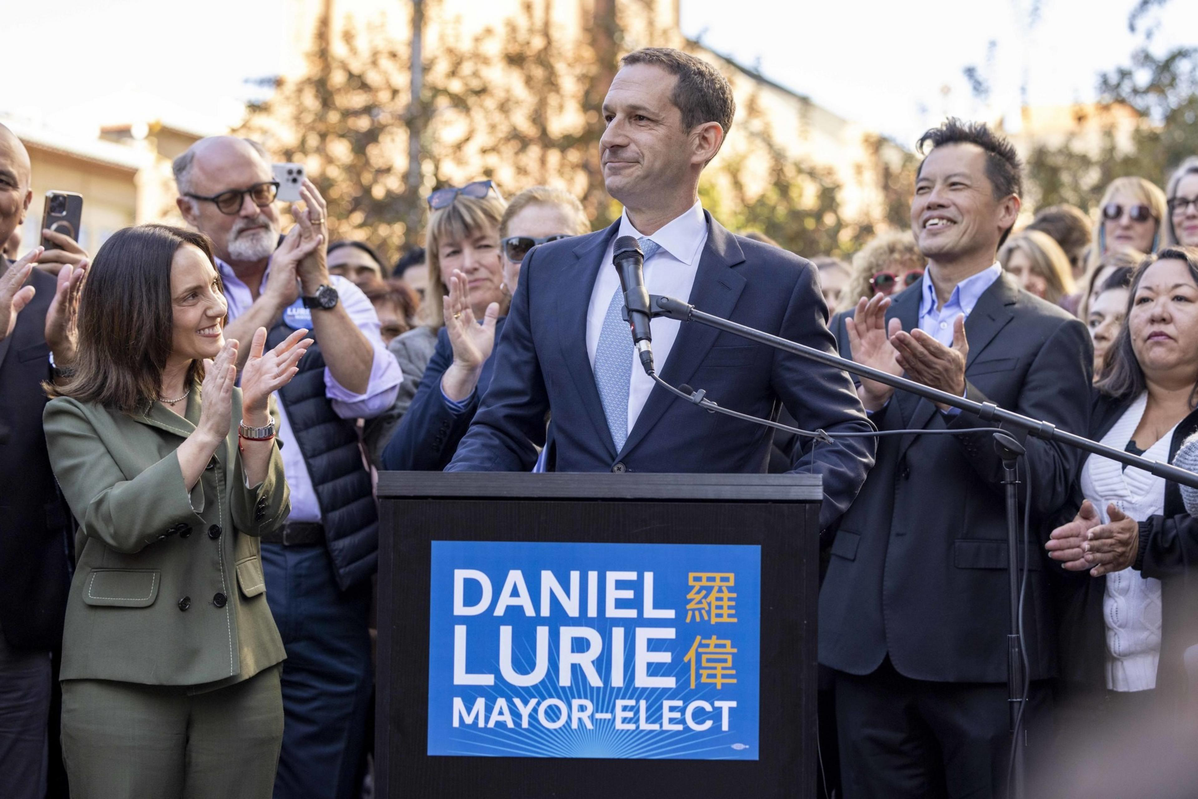 A man stands at a podium labeled &quot;Daniel Lurie Mayor-Elect,&quot; smiling, surrounded by a crowd of applauding people in a sunny outdoor setting.