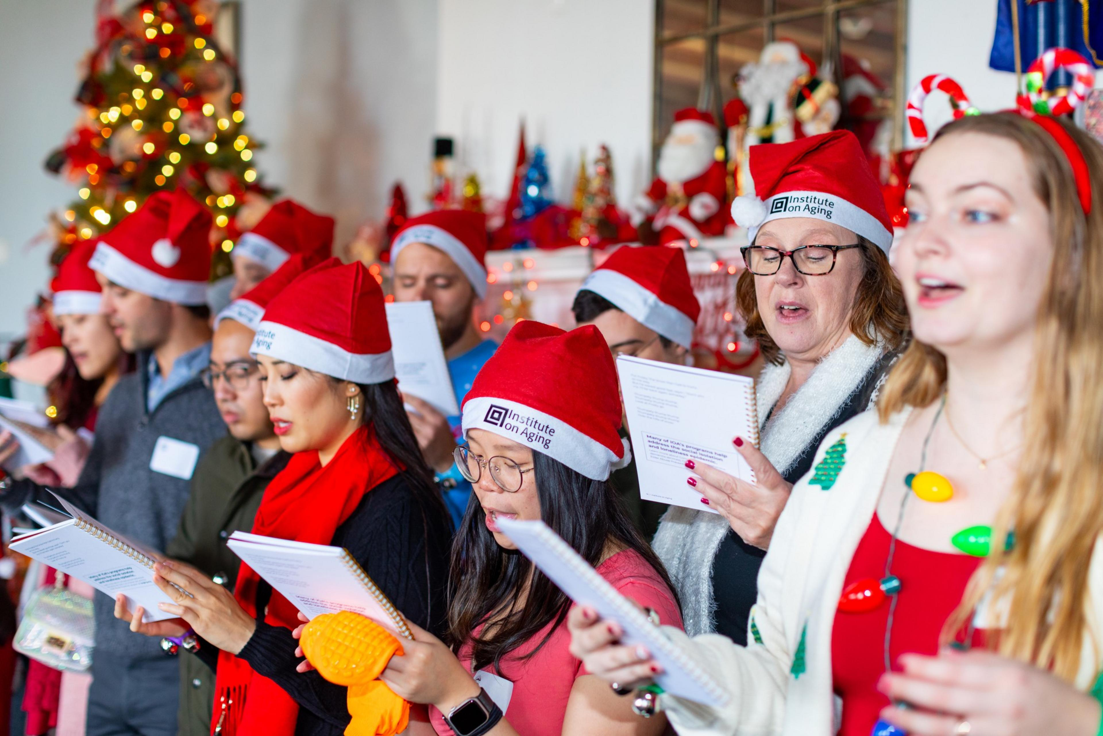 A group of people wearing Santa hats sing Christmas carols, holding songbooks, with a decorated tree and festive lights in the background.