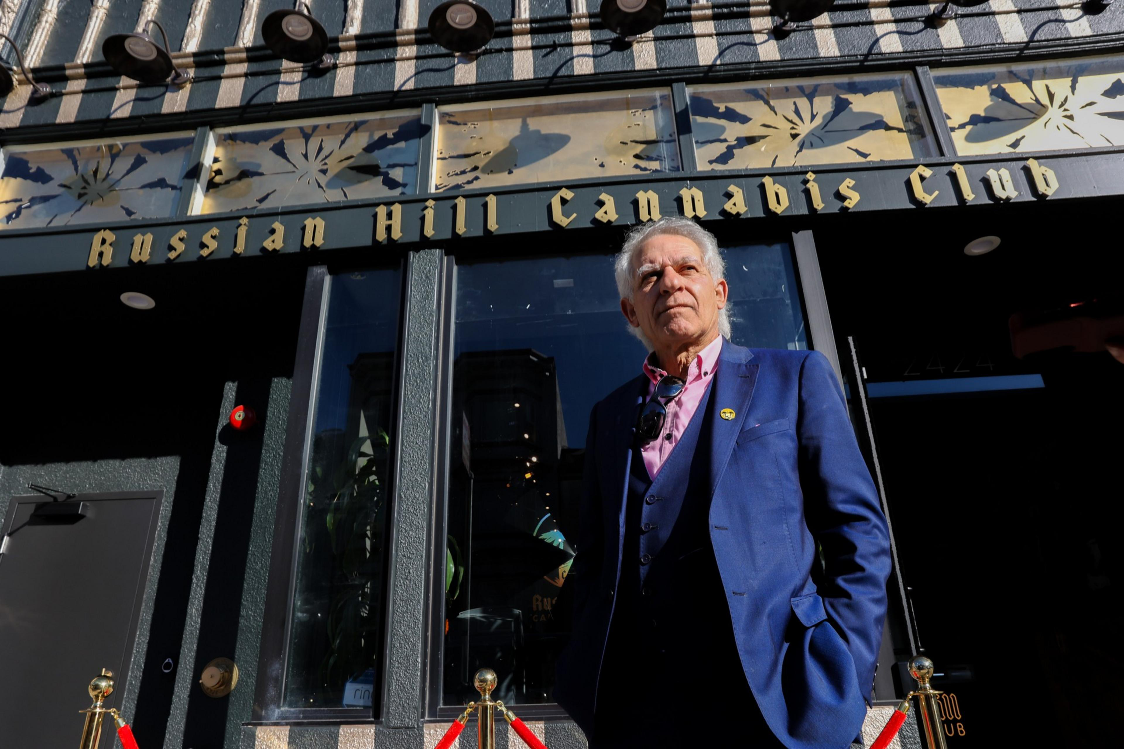 An older man in a blue suit stands confidently outside the Russian Hill Cannabis Club, with sunlight casting shadows on the building facade.