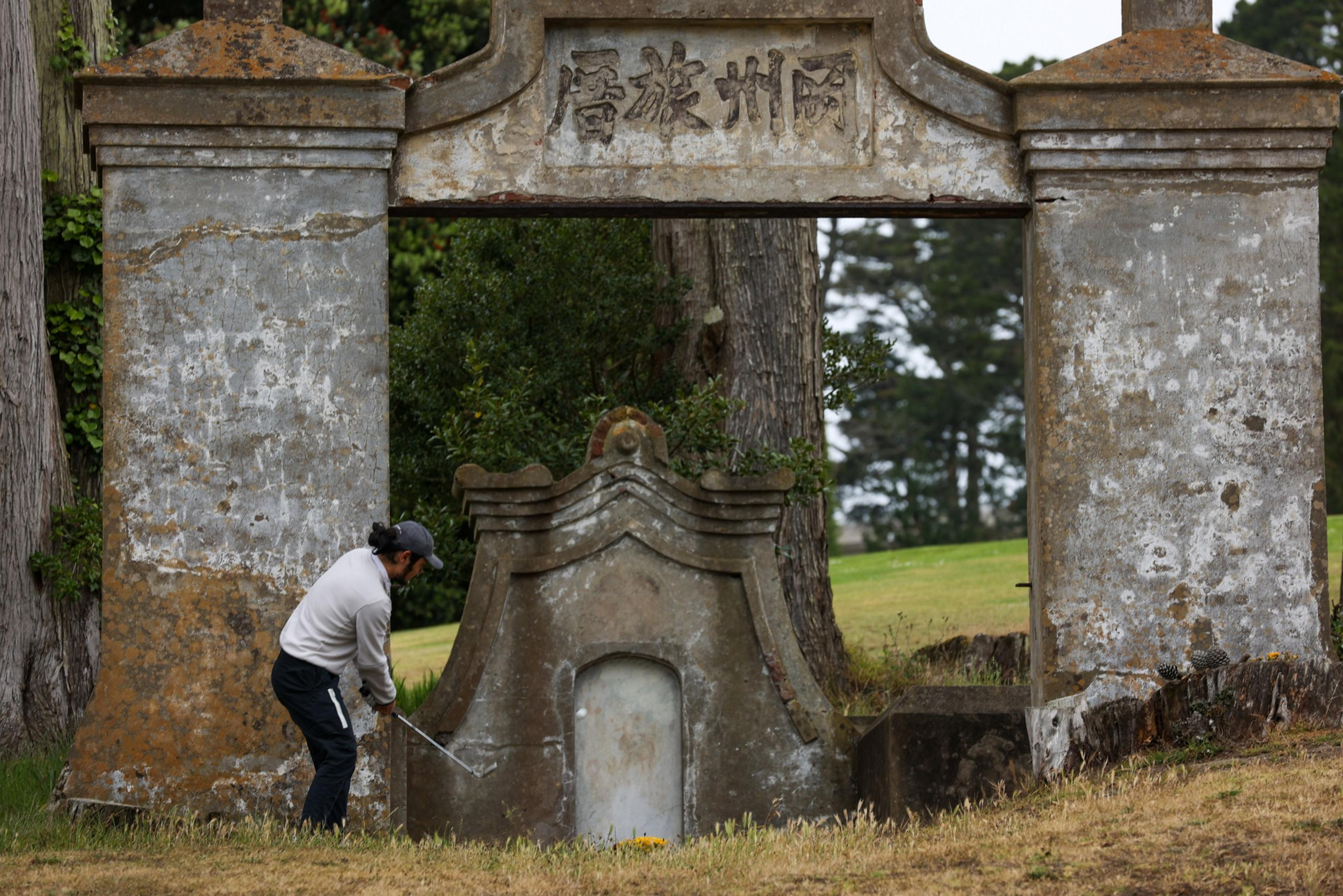 A person wearing a cap swings a golf club near an old stone archway with Chinese characters, surrounded by trees and grass.